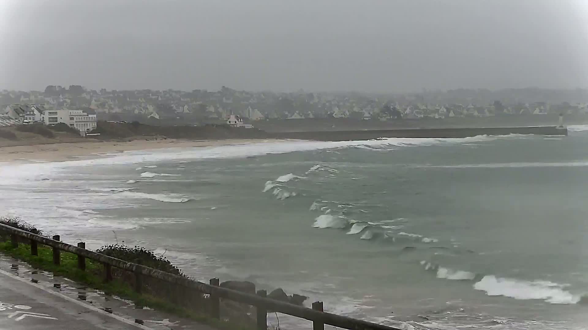 Waves crash on a beach in front of a town under an overcast, gray sky.