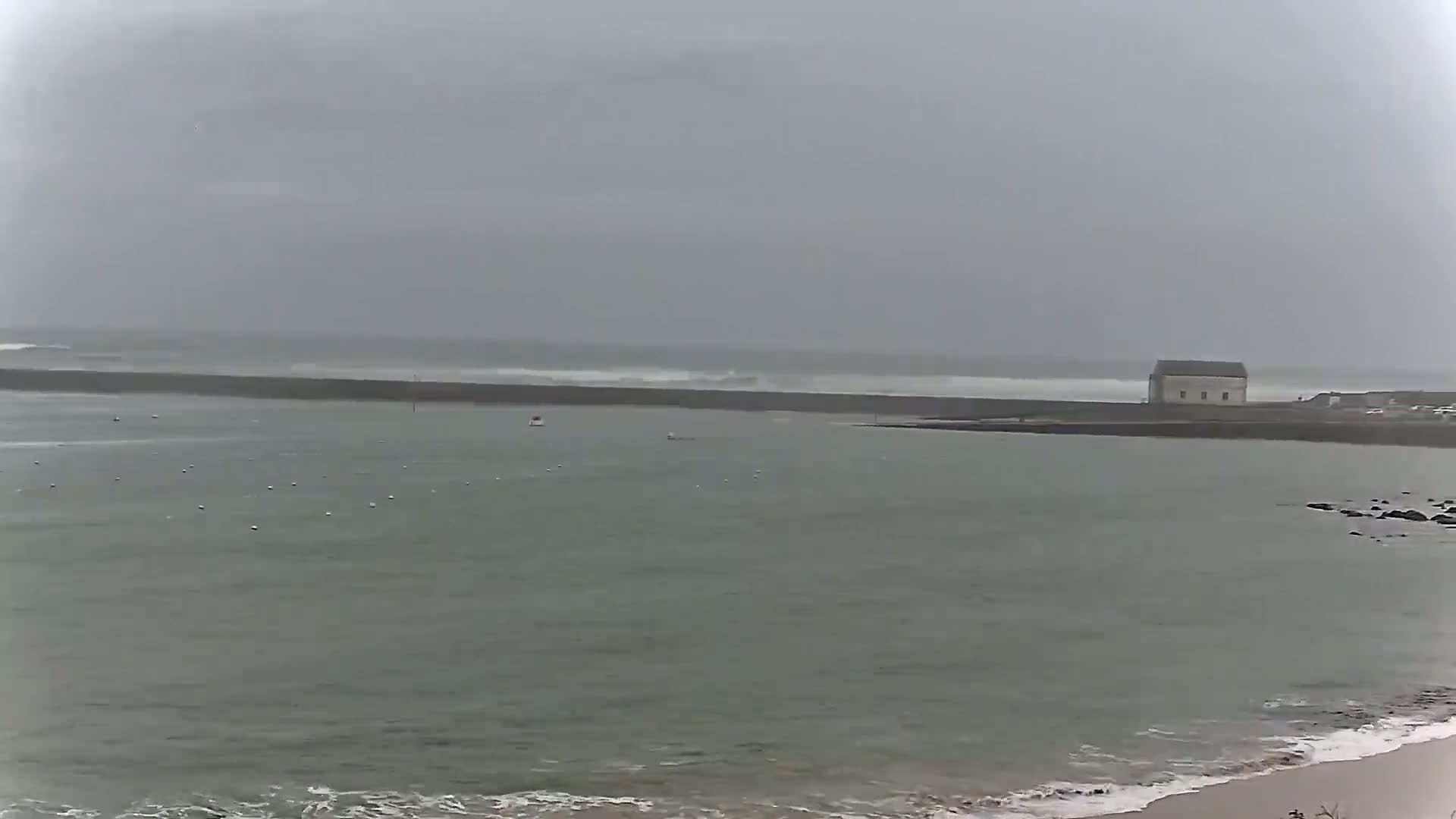 A calm bay with a small building on the far shore is partially protected by a long breakwater against a gray, overcast sky and choppy waves.