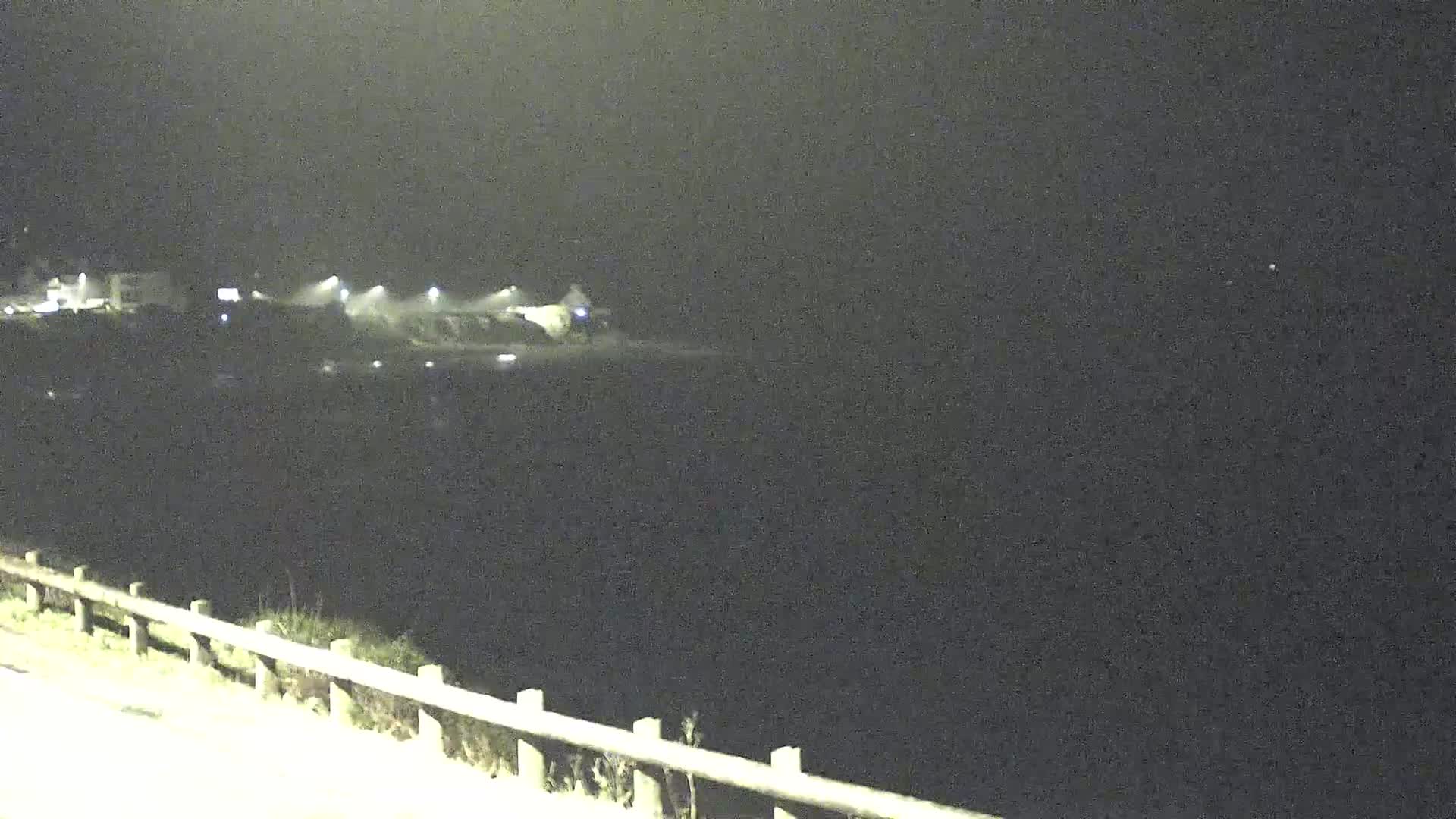 A nighttime, dimly lit view of a dark body of water, with a shoreline featuring some buildings and lights in the distance, viewed from behind a guardrail.