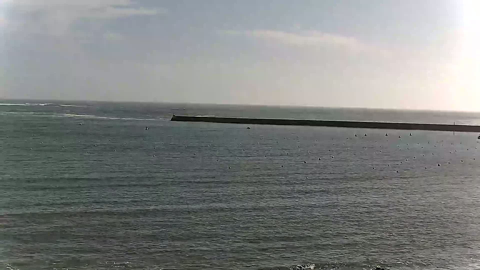 A long, dark breakwater stretches across a calm, gray ocean under a mostly cloudy sky.