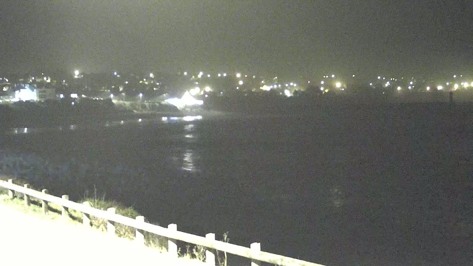 A nighttime view of a dark body of water reflecting distant lights from a town under a cloudy sky.