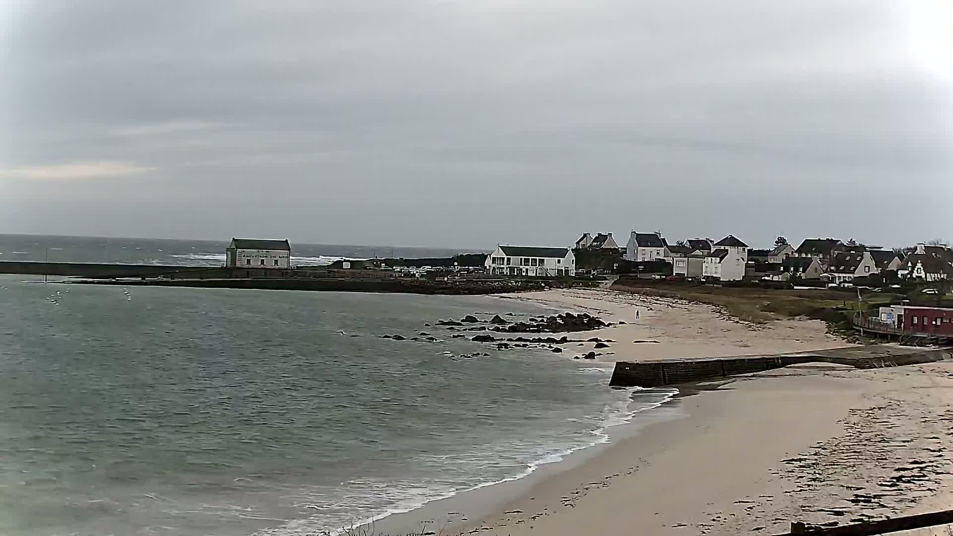 A sandy beach with gentle waves laps against a sea wall, overlooking a small town under a cloudy sky with a choppy ocean in the distance.