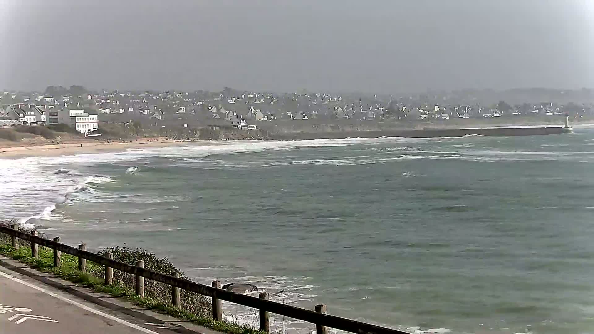 A grey, overcast day shows a view of a beach with moderate waves and a town in the distance.