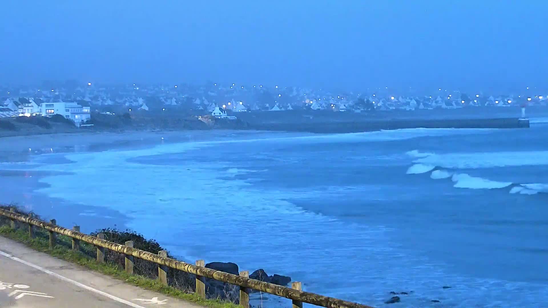 A coastal town is visible in the distance under a twilight sky, with moderately sized waves rolling onto a beach in the foreground.