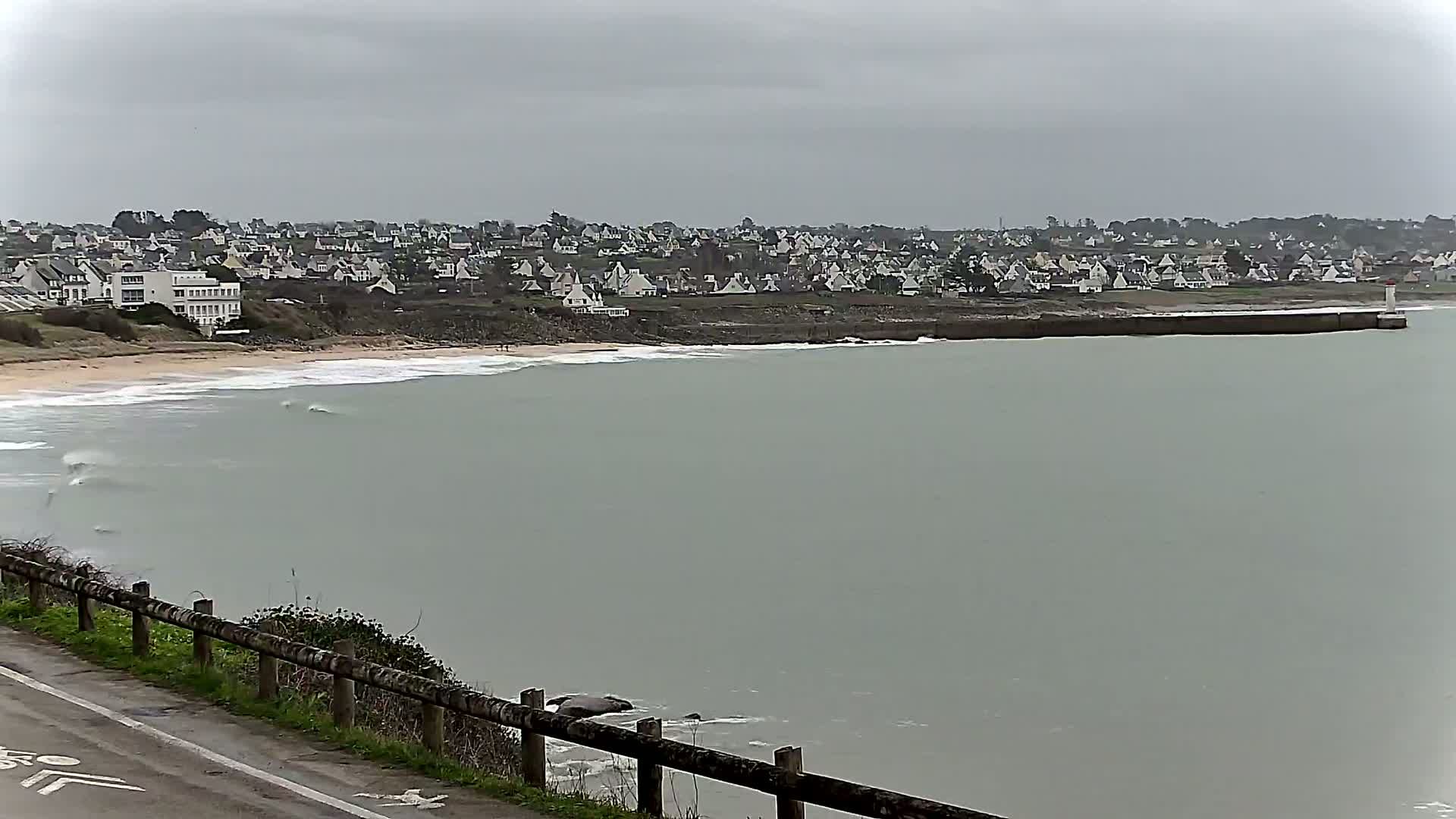 A coastal town with white houses is seen across a calm, gray sea under an overcast sky, from a viewpoint above a sandy beach and a wooden fence bordering a road.