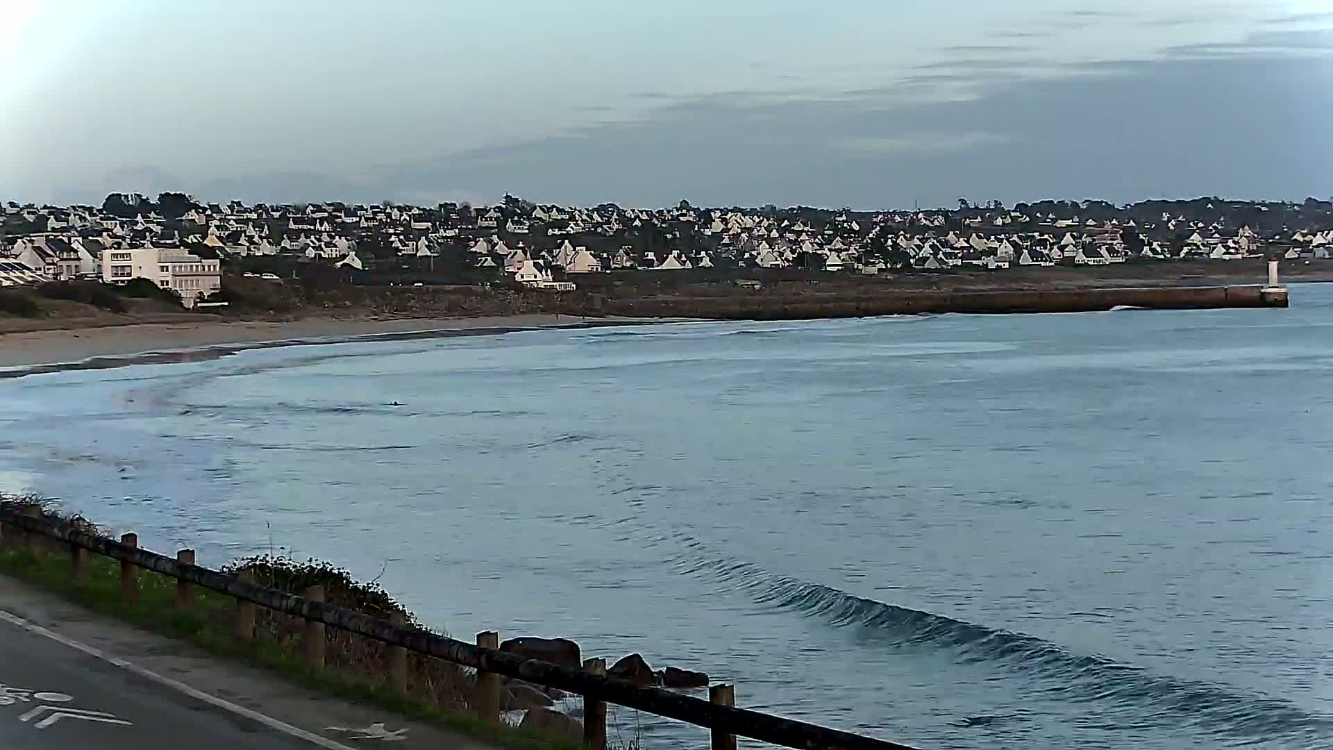 A coastal town sits atop a small cliff overlooking a calm ocean with gentle waves rolling onto a sandy beach.
