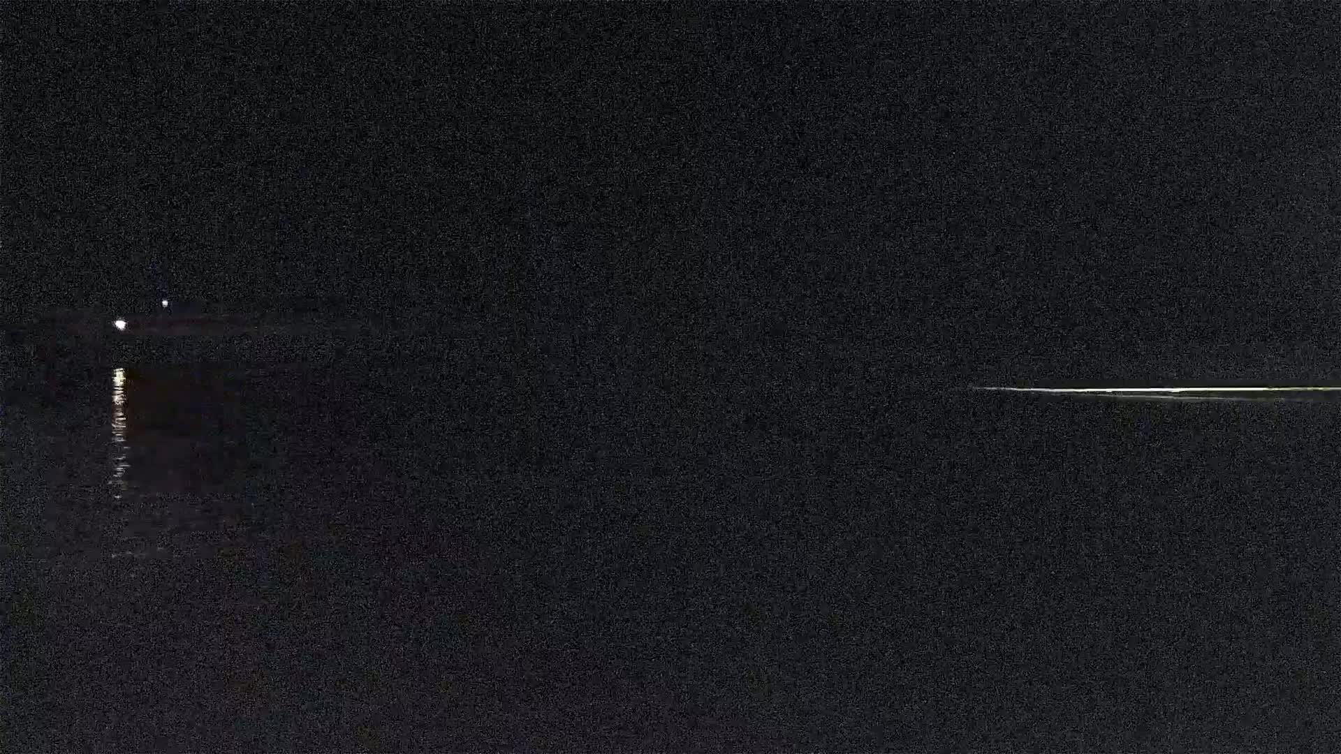 A dark, nighttime scene shows a body of water reflecting faint lights from a distant shore.