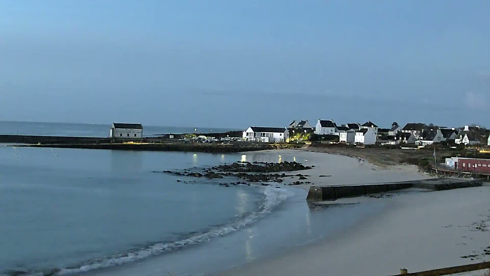A small coastal town with white buildings is nestled beside a calm, light-blue ocean under a twilight sky.