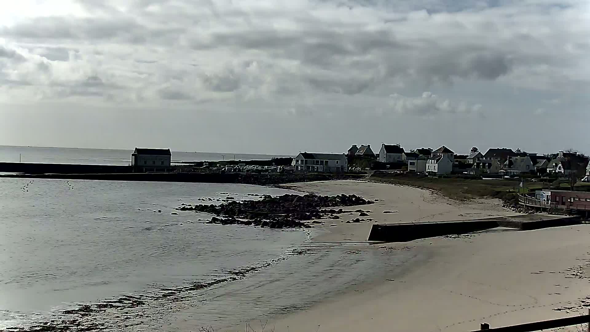 A sandy beach with calm water and a small village in the background under a cloudy sky.
