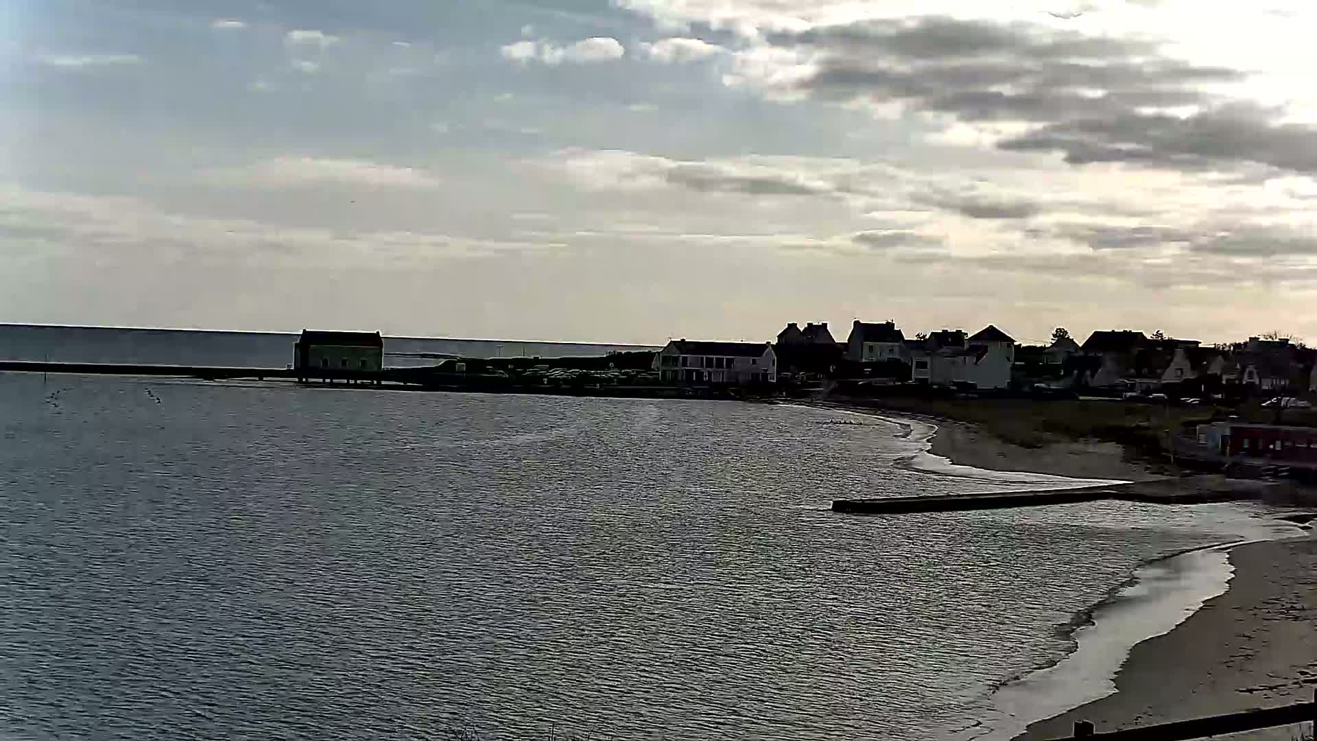A calm body of water laps a sandy beach near a row of houses under a cloudy sky.