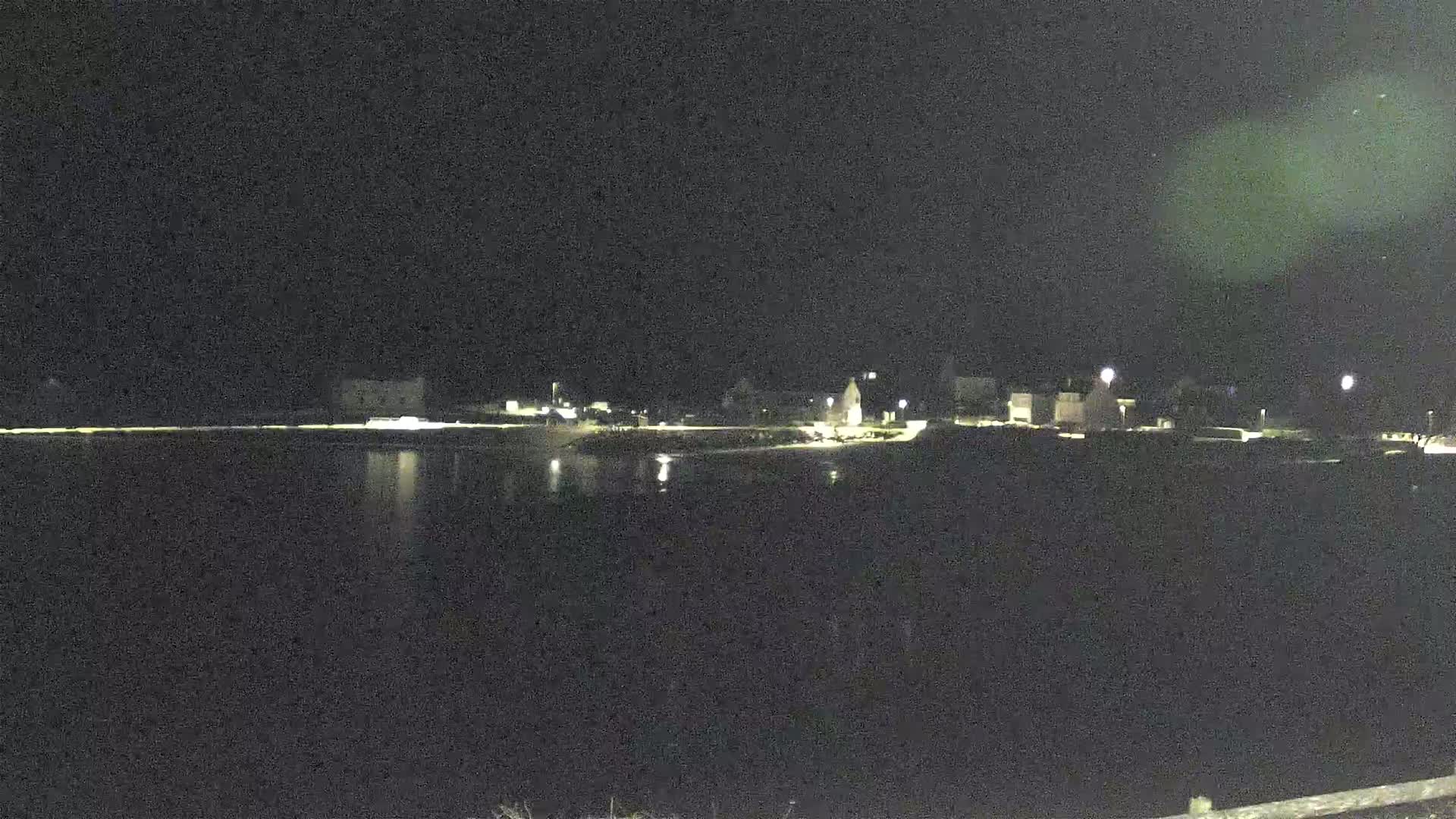 A dark, nighttime scene shows a body of water reflecting the faint lights of a small town across the shore.