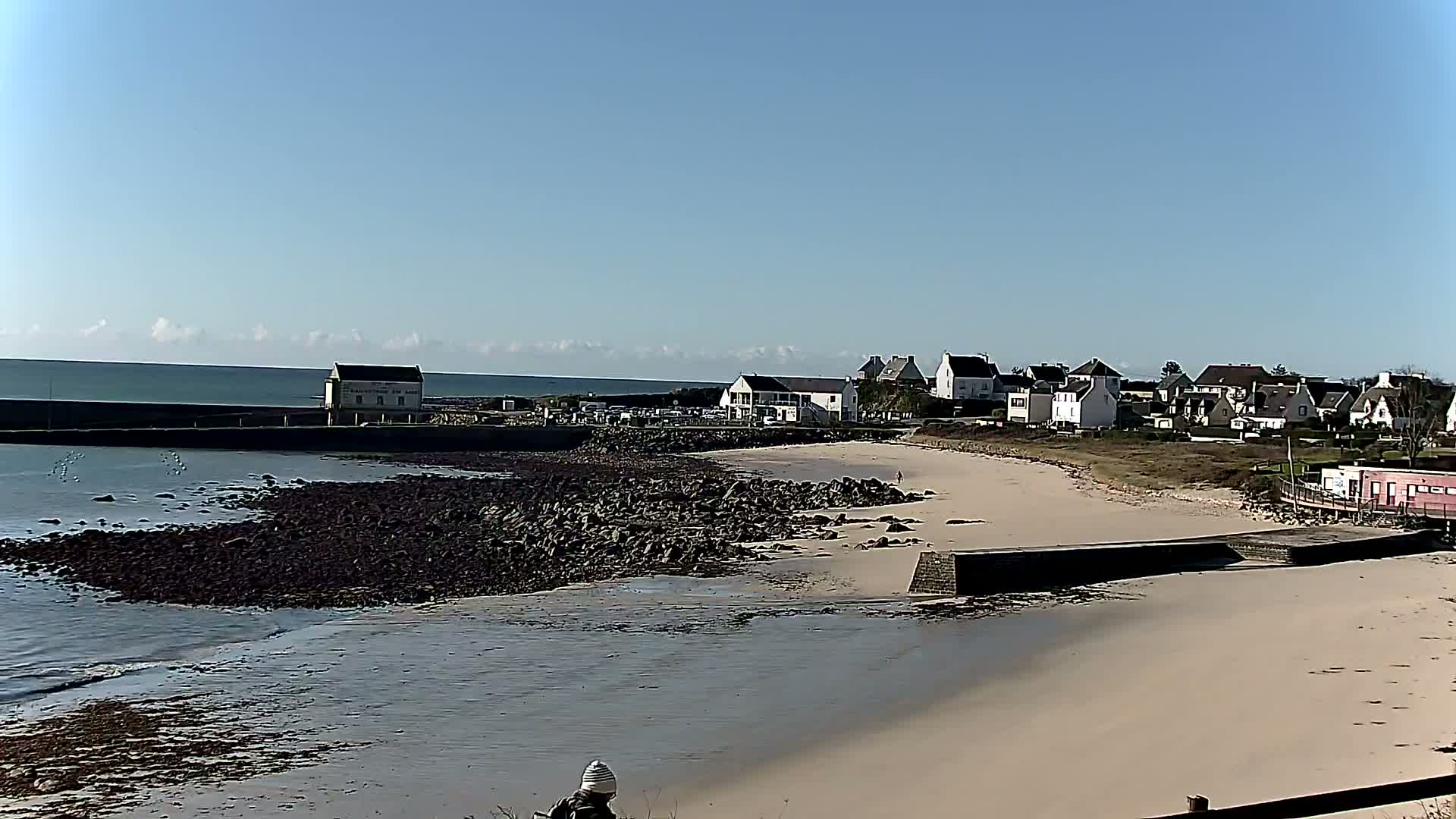 A sandy beach with dark rocks and a small town beyond it under a clear blue sky.