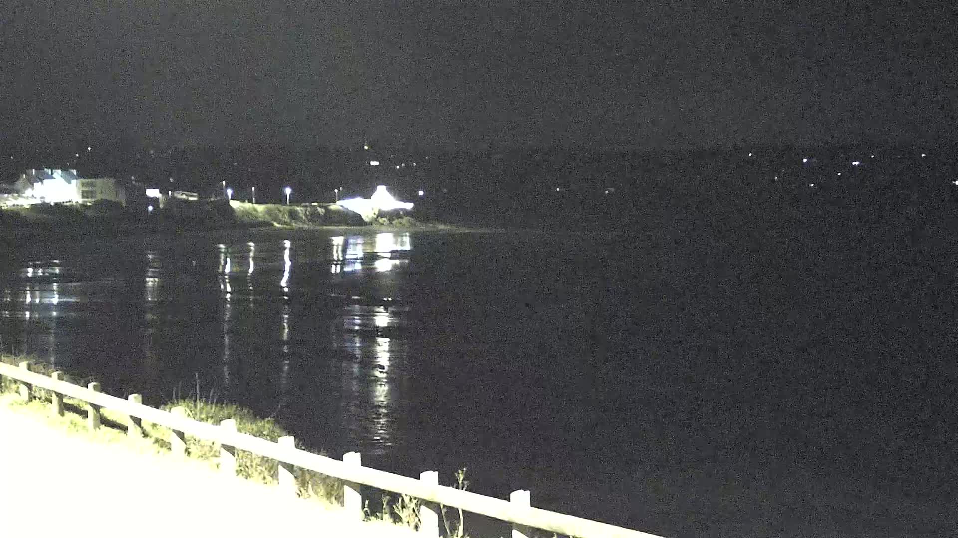 A nighttime view of a calm body of water reflecting lights from a distant shoreline, with a low fence and road in the foreground.