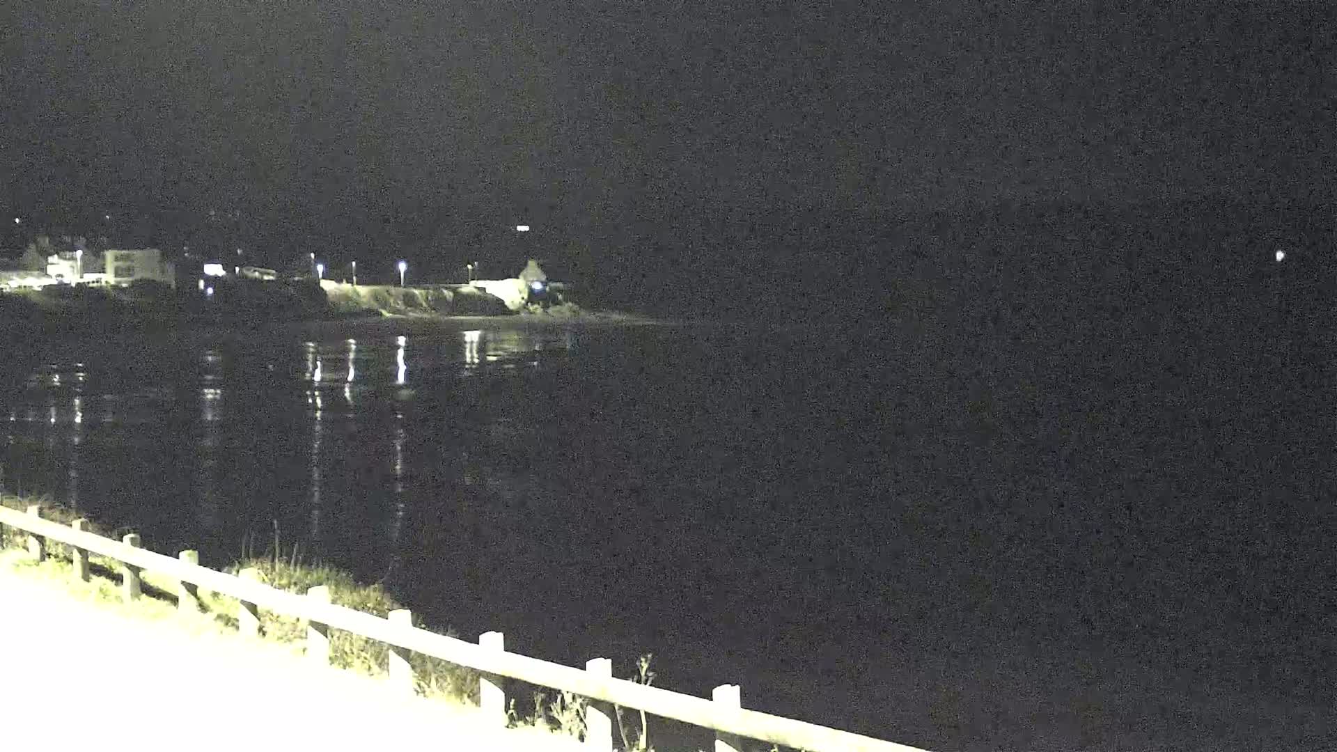 A nighttime view shows a dark body of water reflecting lights from a shoreline with buildings and a few streetlights.