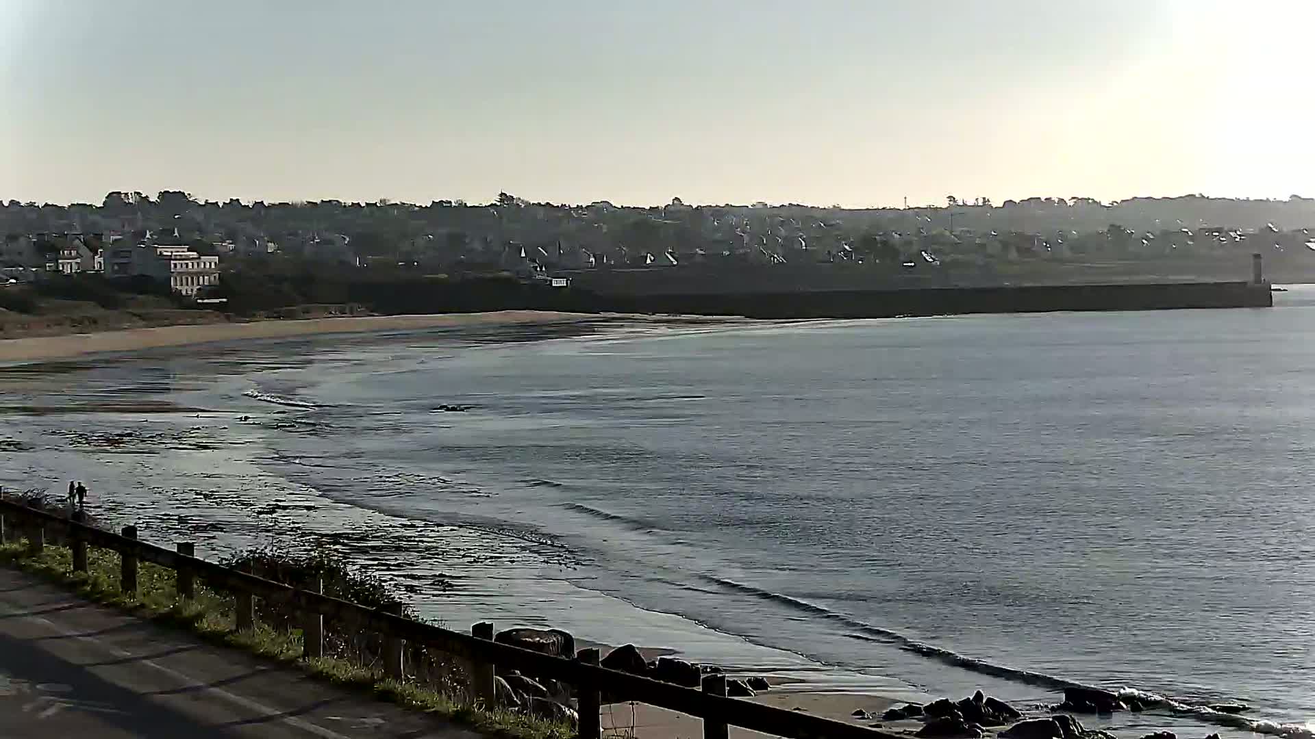 A sandy beach with gentle waves laps the shore under a clear, bright sky, with a town and a breakwater visible in the distance.