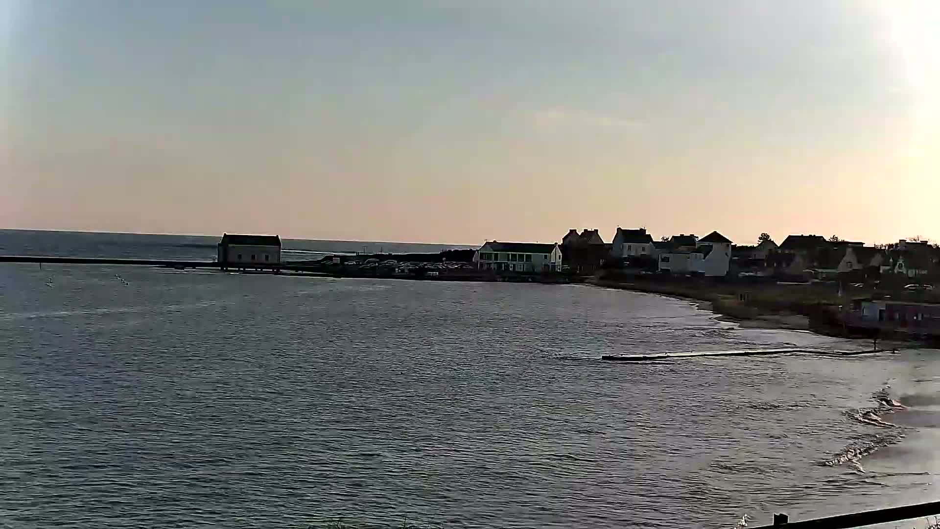 A calm body of water laps gently against a sandy shore where a small village of houses sits under a clear, pale sky.