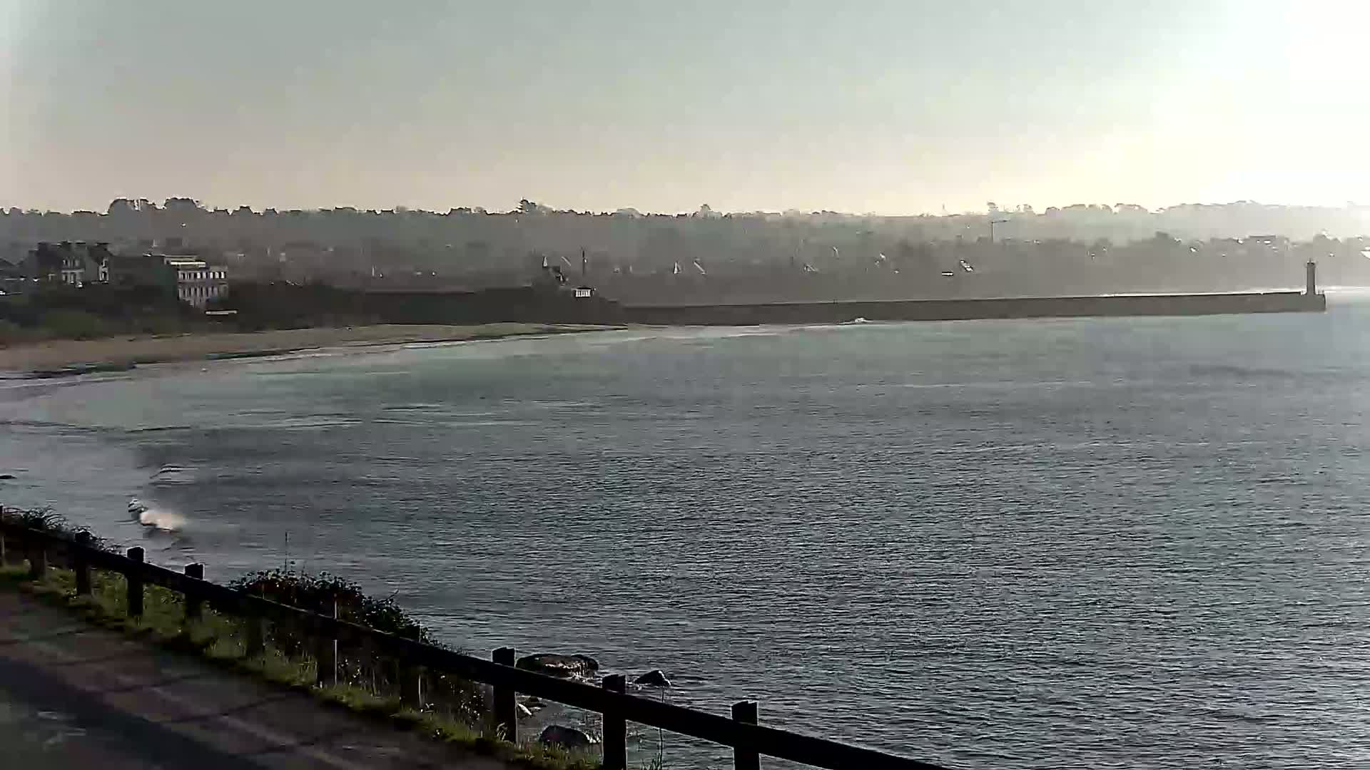 A hazy, overcast day reveals a calm ocean with small waves lapping at a sandy beach, backed by a long seawall and a distant town.