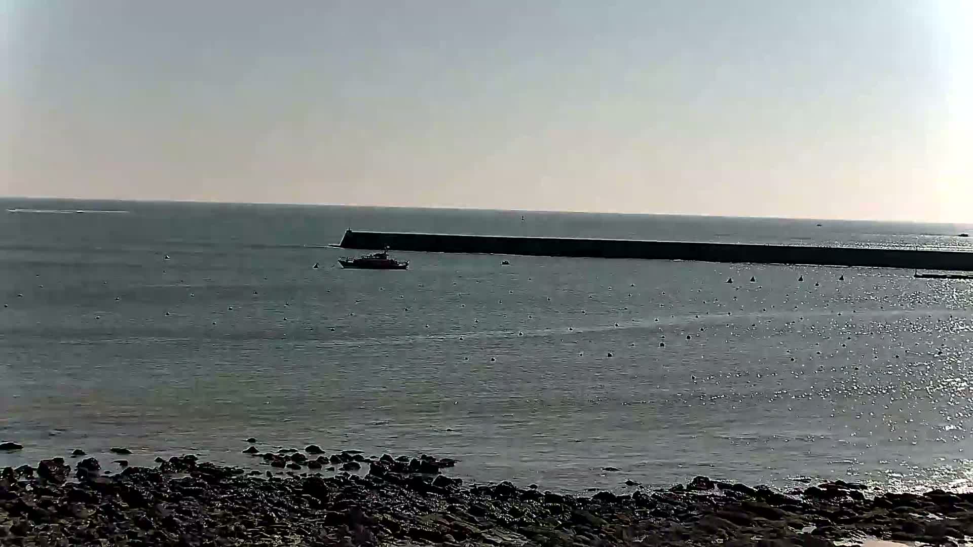 A calm, sunny day shows a long breakwater stretching across a gray sea, with a small boat in the foreground and a rocky shore in the bottom of the image.
