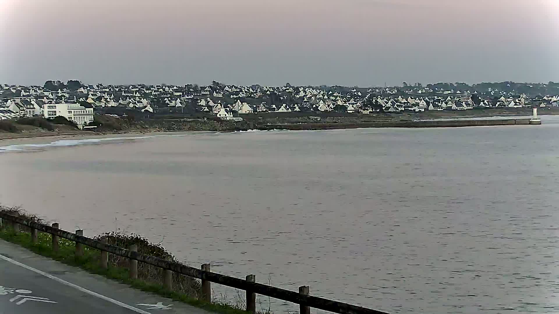 A coastal town is visible across a calm, grey body of water from a roadside vantage point.