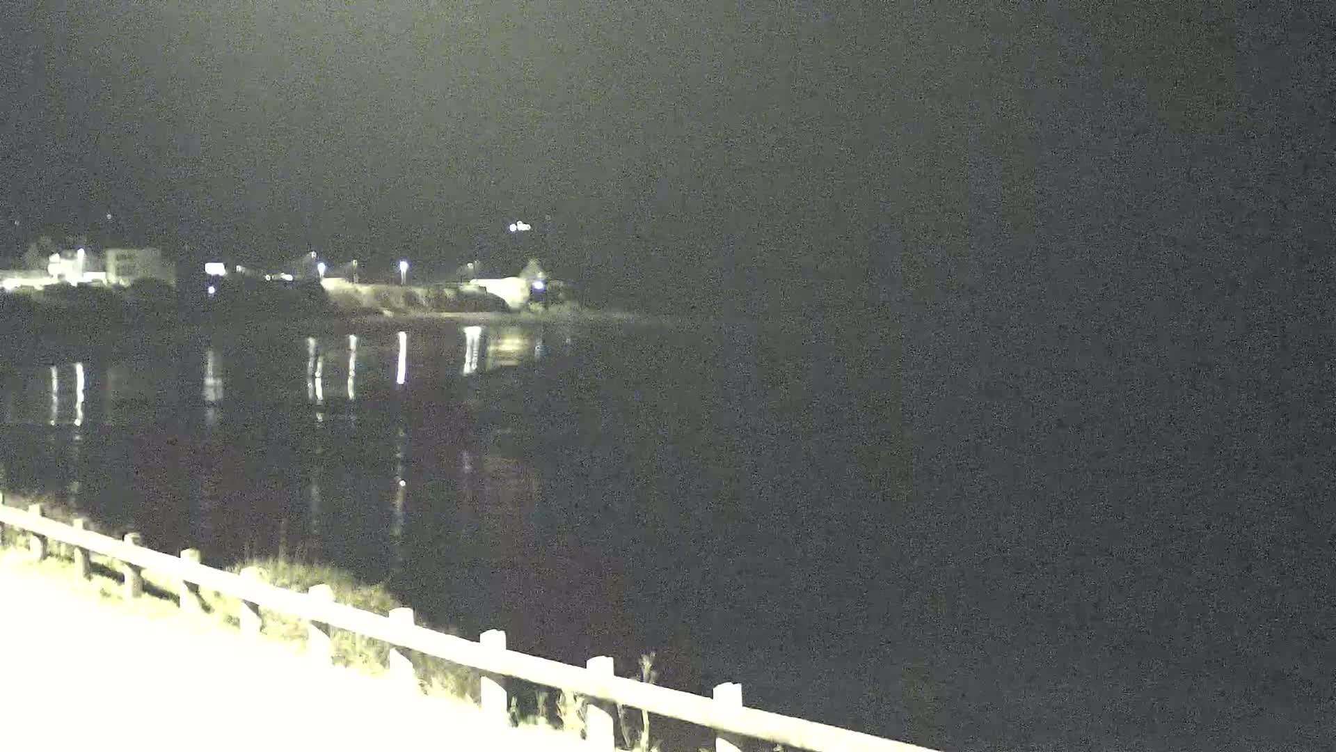 A nighttime view of a dark body of water reflecting faint lights from buildings across the shore, seen from a slightly elevated vantage point with a low fence in the foreground.