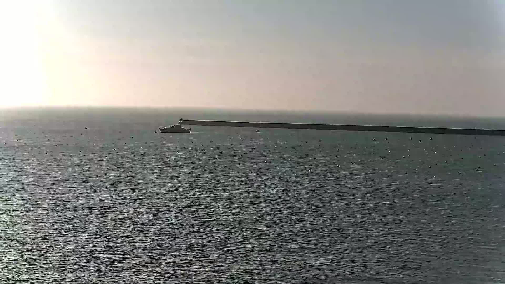 A boat is in calm, gray water in front of a long, dark breakwater under a hazy sky.