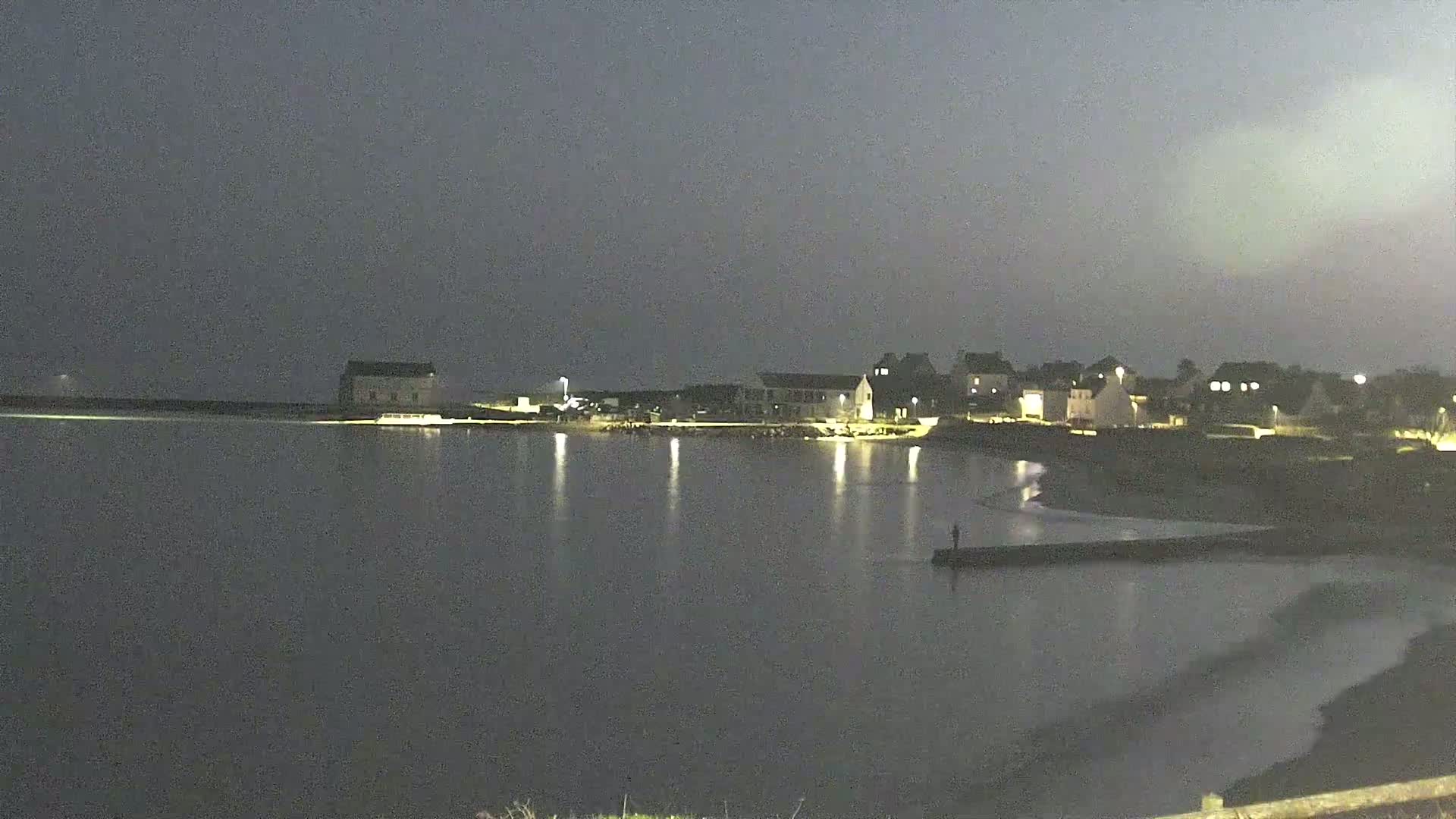 A calm, dark night scene shows a body of water reflecting the lights of a small town on the shore, with a lone figure standing on a jetty.