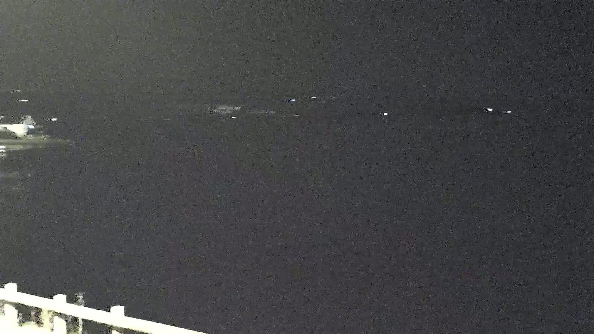At night, a dark body of water is visible, with faintly lit objects possibly on the far shore and a section of a railing or fence in the lower left corner.