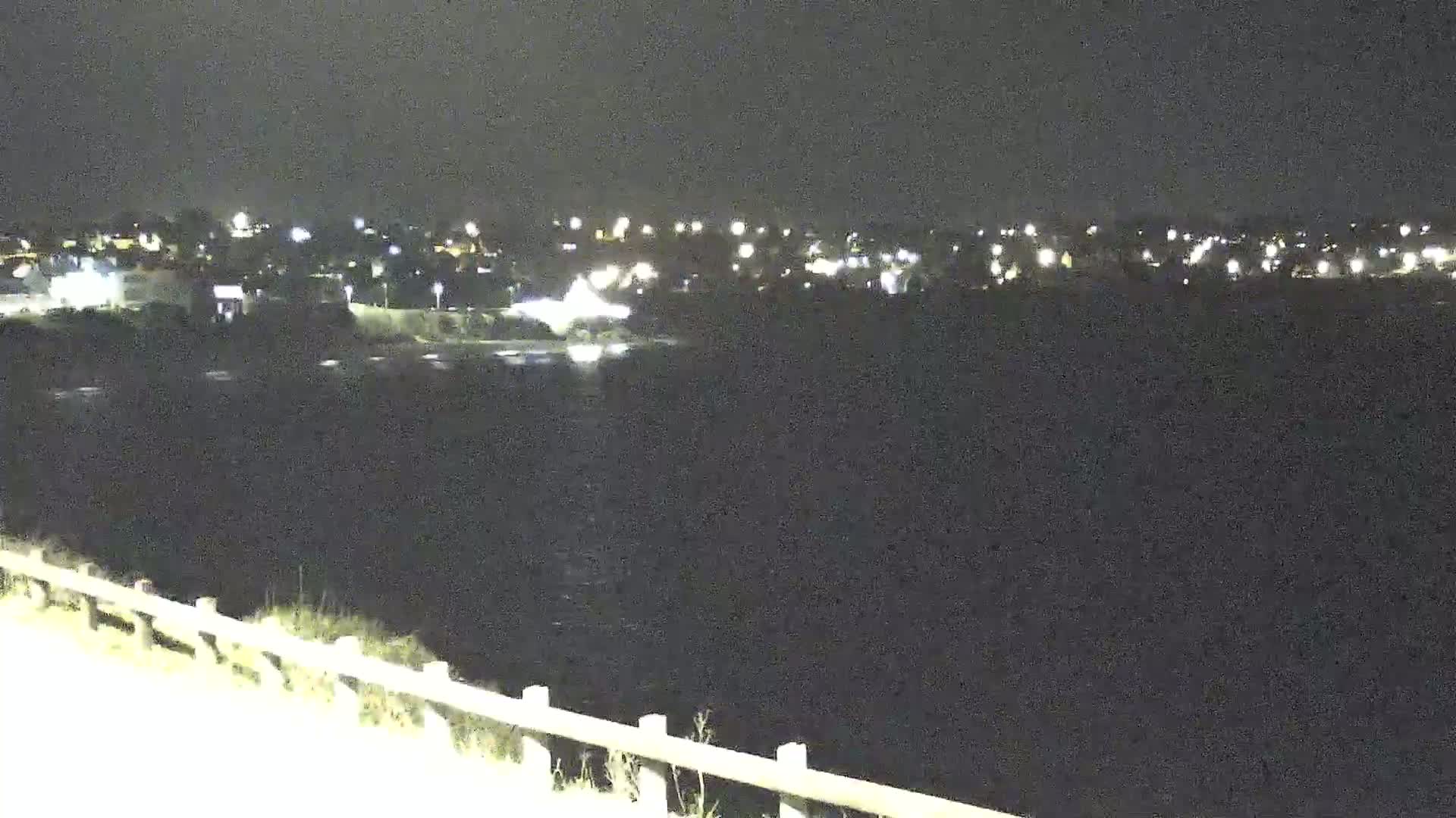 A nighttime view of a dark body of water with a distant town illuminated by lights across the water, seen from a slightly elevated vantage point with a low fence in the foreground.