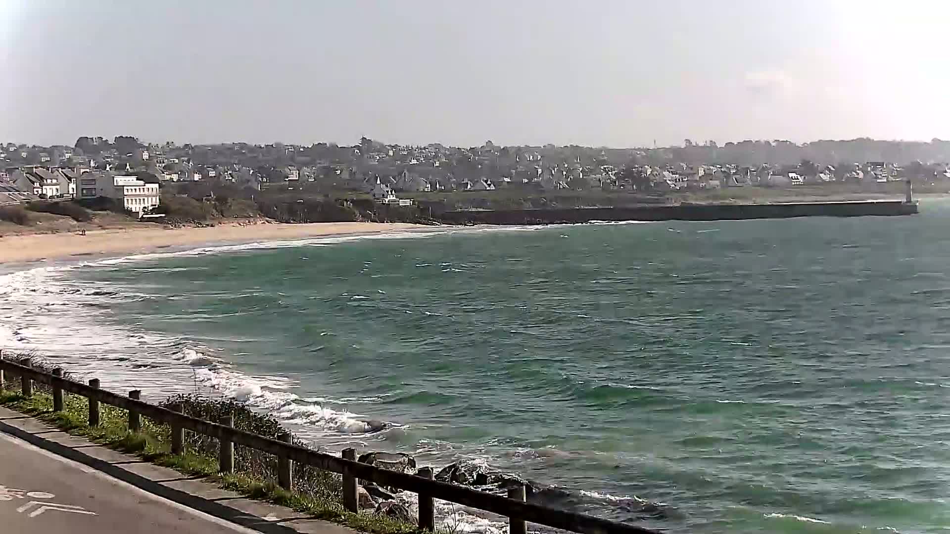 A sandy beach with small waves laps the shore near a town under a bright, mostly clear sky.