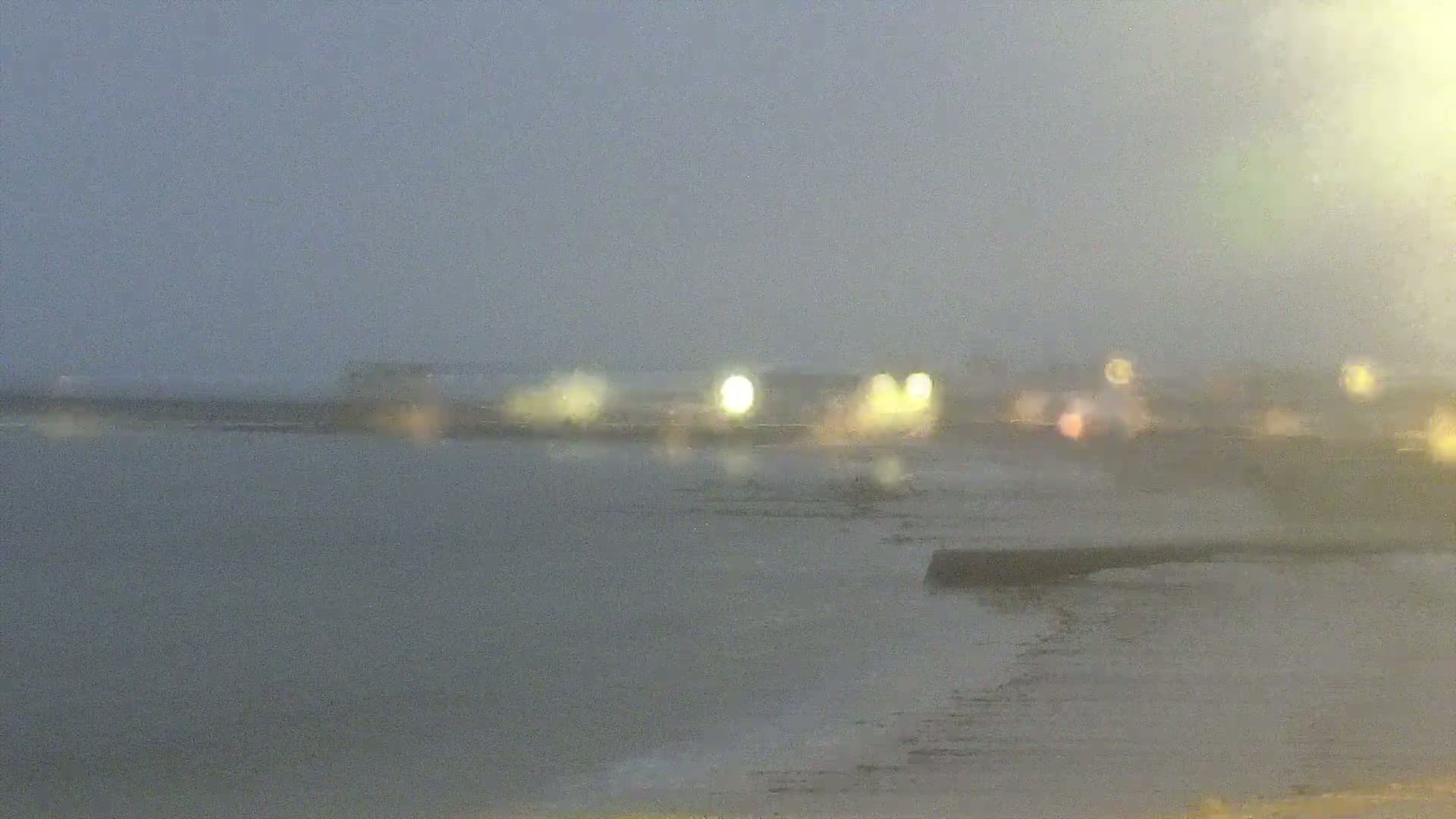A blurry, nighttime coastal scene shows a body of water with a distant, indistinct shoreline of lights under a hazy, possibly rainy sky.