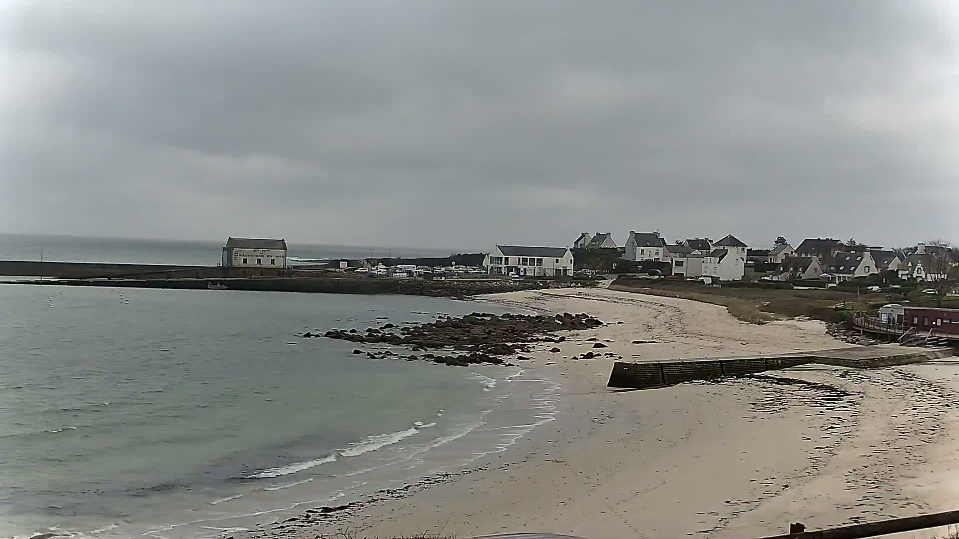 A sandy beach with small waves laps against a rocky shoreline, next to a small town under an overcast sky.