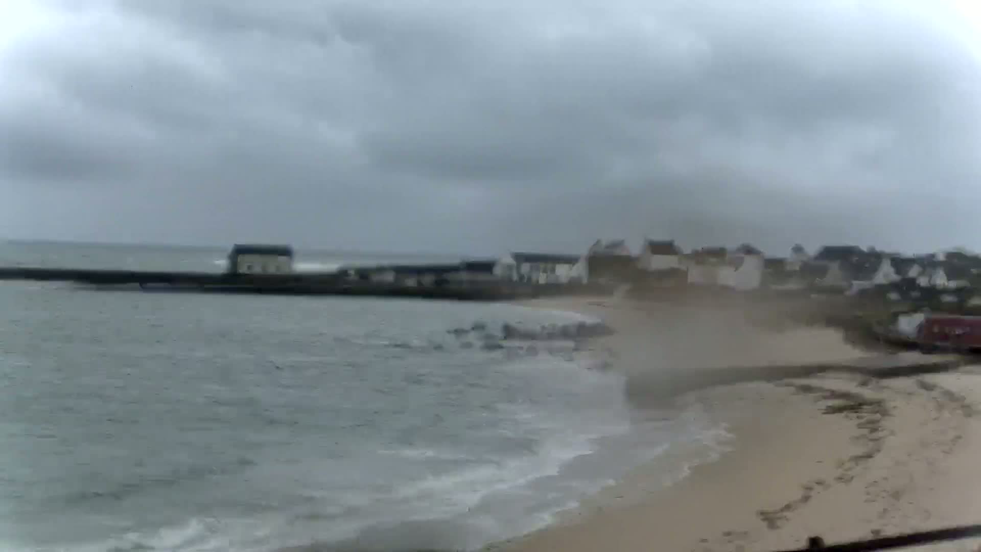A sandy beach with small buildings and a seawall is shown under an overcast sky with choppy water.