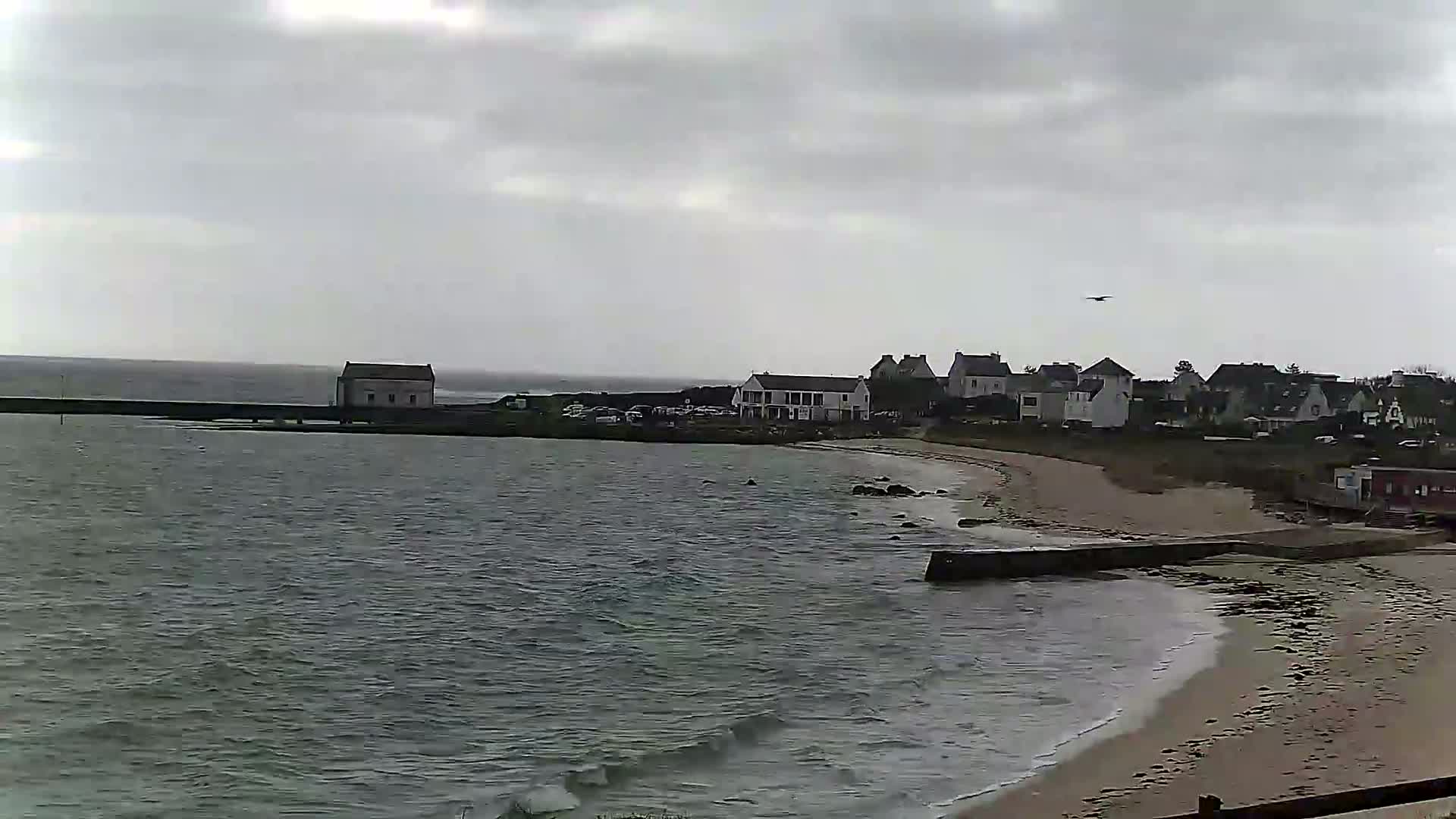 A grey, overcast day reveals a coastal town with a small sandy beach and calm, grey water.