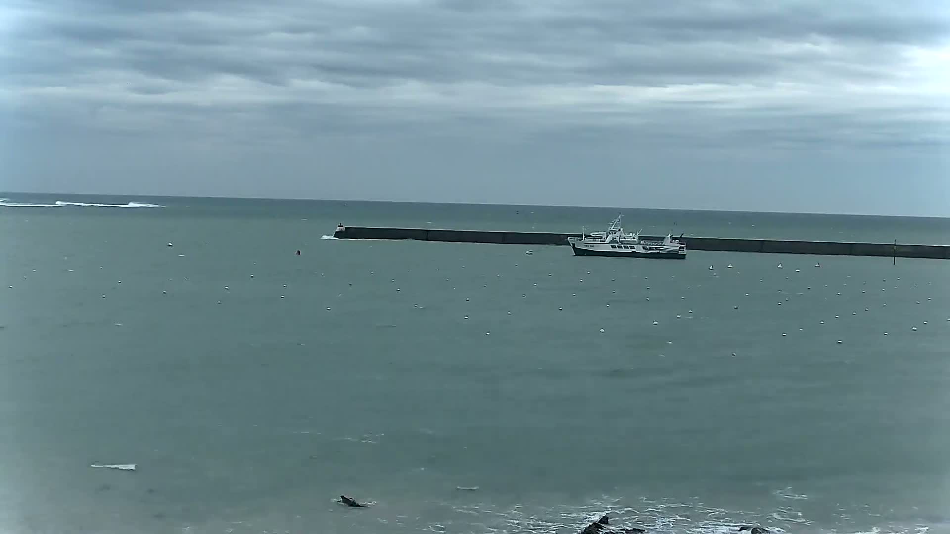A ferry is moored near a long breakwater in a calm, gray-sky sea with small waves.