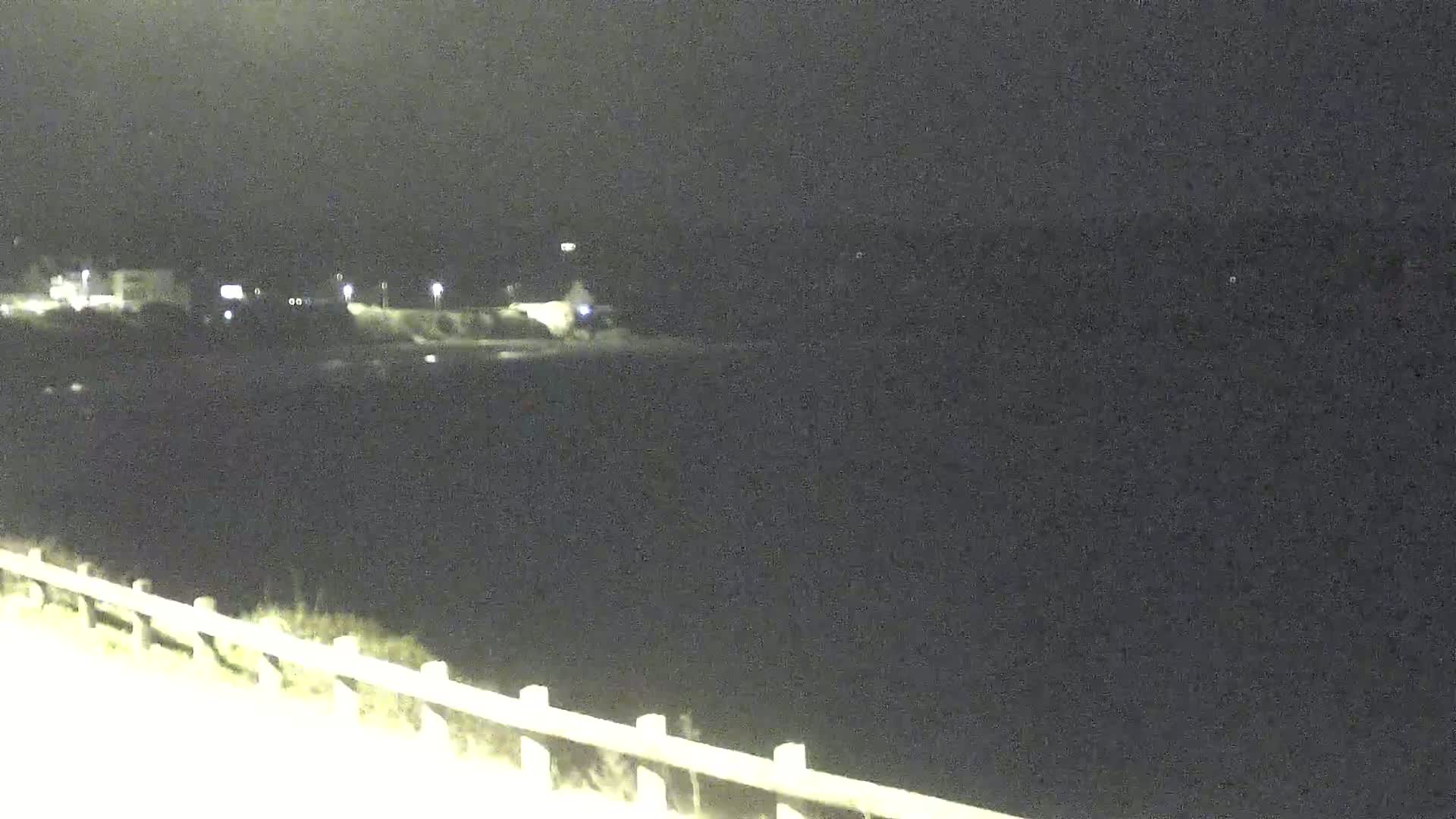 A nighttime view shows a dark body of water, a distant shoreline with faint lights, and a low fence or railing in the foreground.