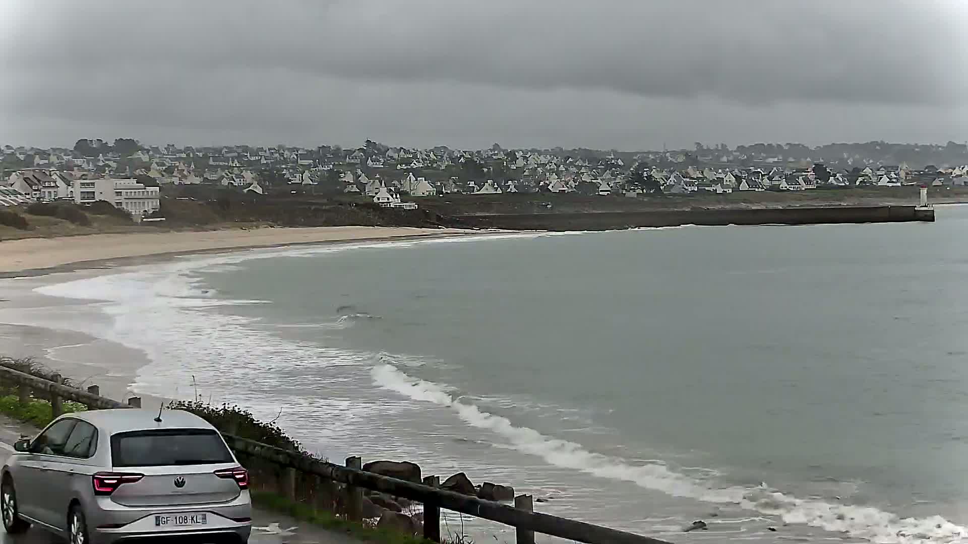 A silver car is parked near a beach with small waves rolling onto the shore under an overcast sky, with a town visible in the distance.