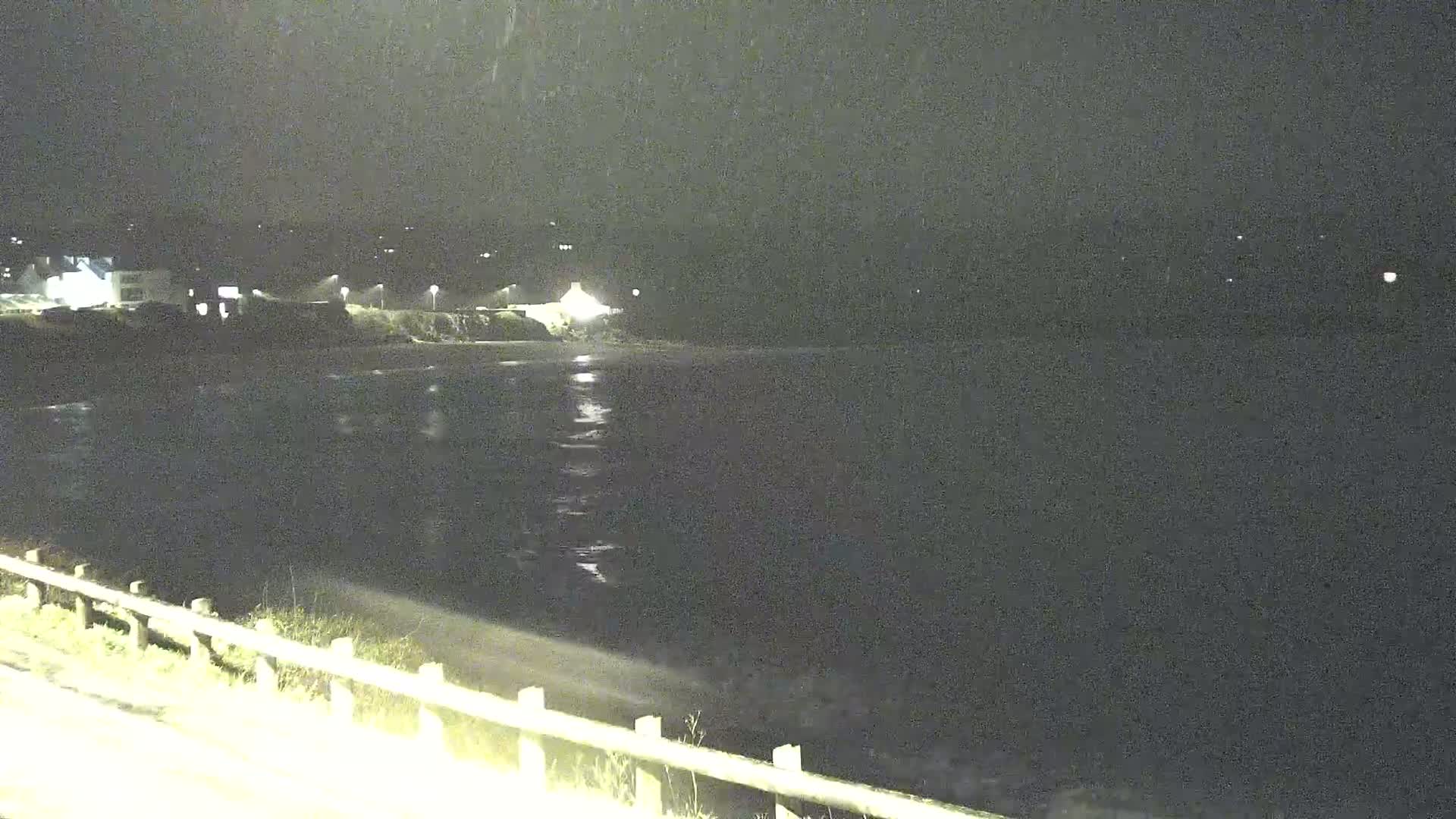 A nighttime view of a dark body of water under a lightly snowing sky, with a shoreline featuring buildings and a low fence in the foreground.