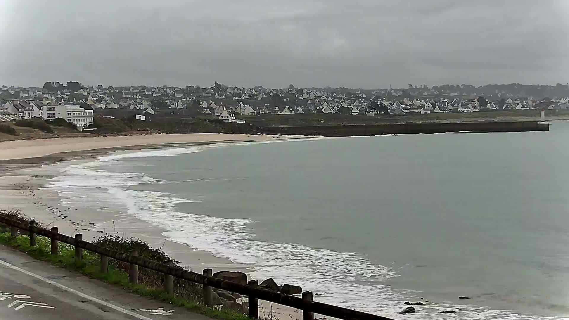 A sandy beach with small waves rolls onto the shore near a town of white houses under a gray, overcast sky.