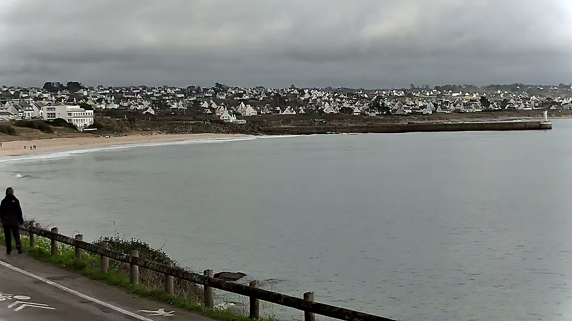 A person walks along a road overlooking a calm, gray sea with a sandy beach and a town of white houses under an overcast sky.