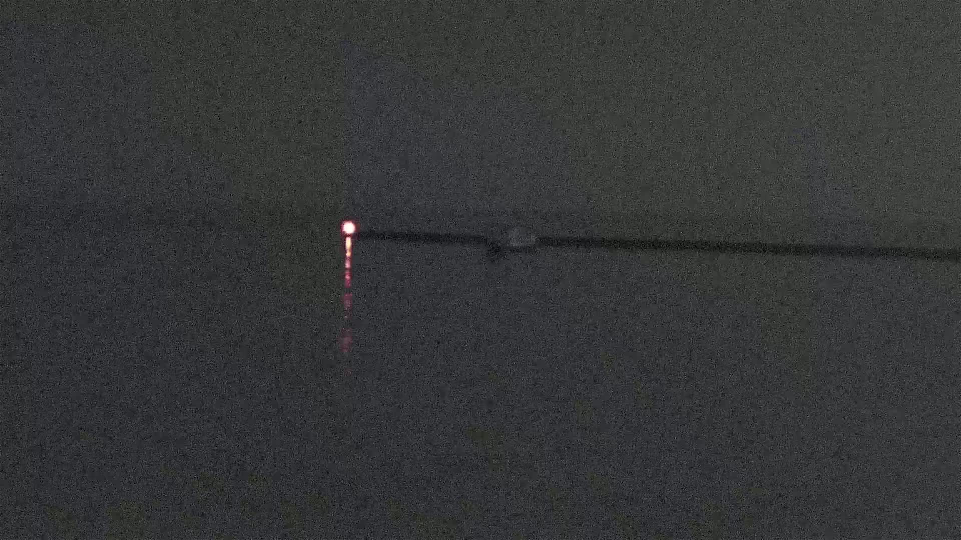 A dark, calm body of water reflects a single, small, red light at night.