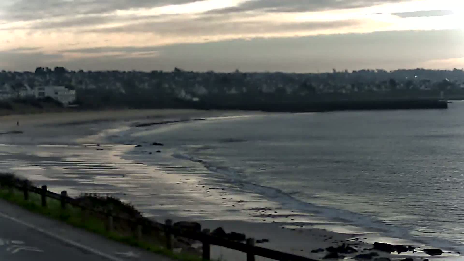 A sandy beach with gentle waves curves along a coastline under a cloudy, overcast sky, with a residential area visible in the background.