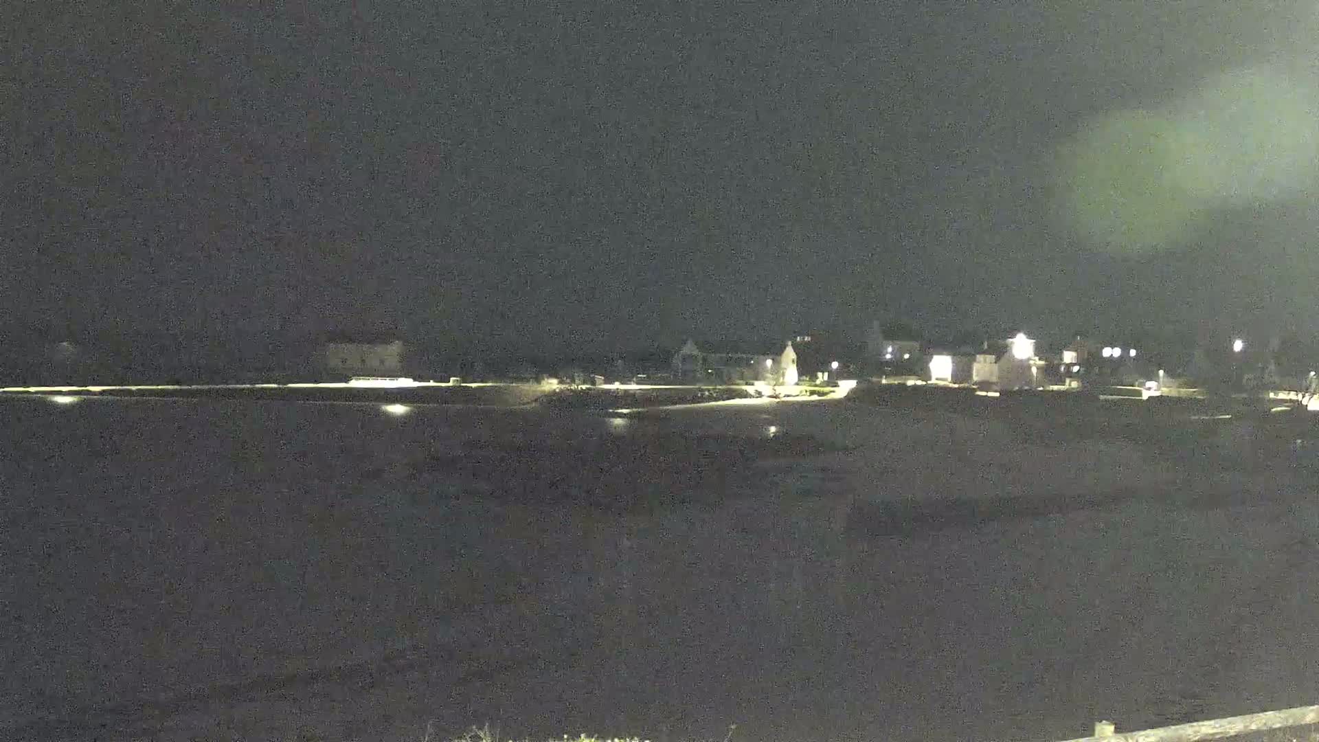 A nighttime view of a sparsely lit town across a dark body of water under a cloudy sky.