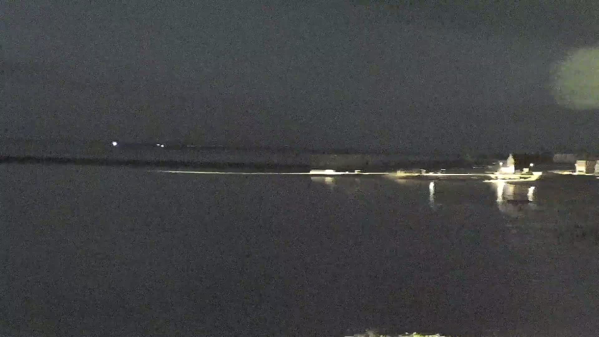 A nighttime view shows a dark body of water reflecting the lights of a distant shoreline.