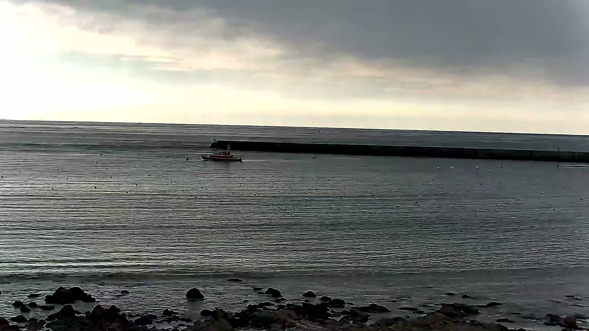 A small boat is on calm, gray water near a long breakwater under an overcast sky.