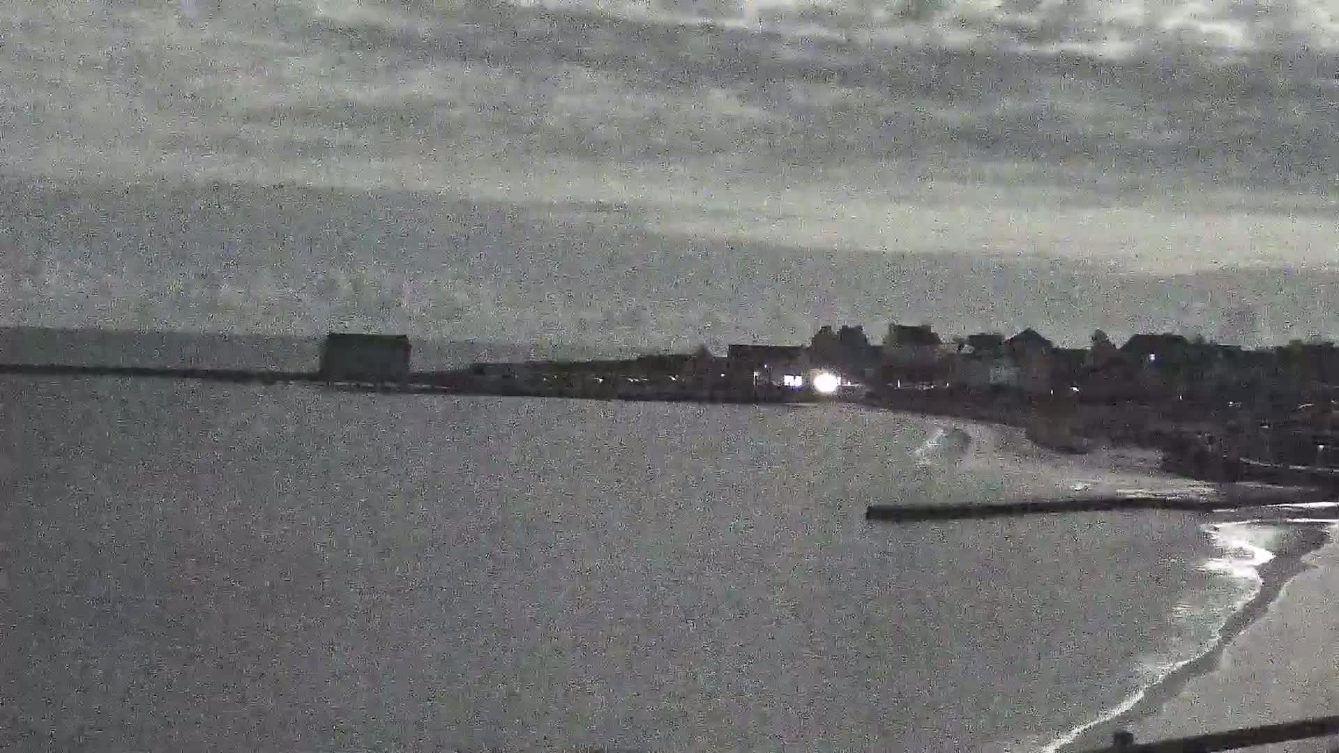 A dark, overcast sky looms over a calm body of water lapping a shoreline with buildings and a breakwater.