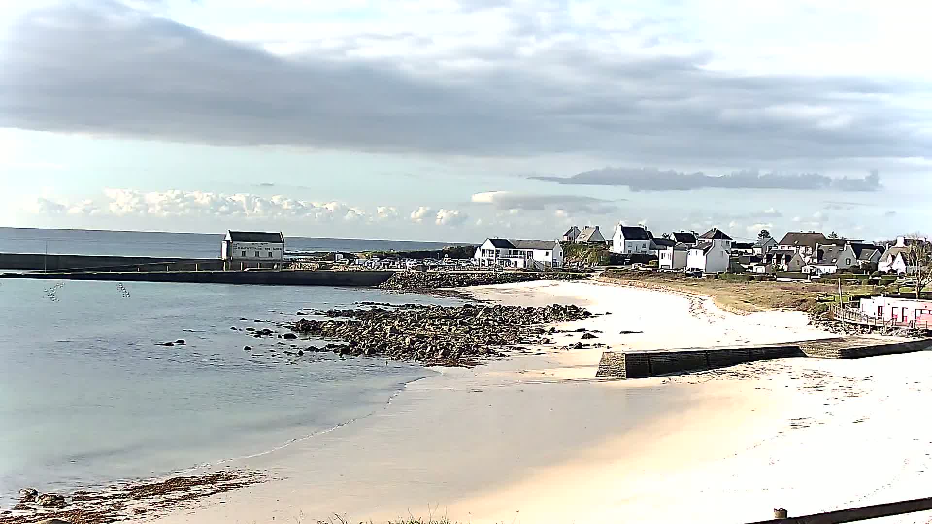 A sandy beach curves around a rocky shoreline, with a small town and calm ocean visible in the background under a cloudy sky.