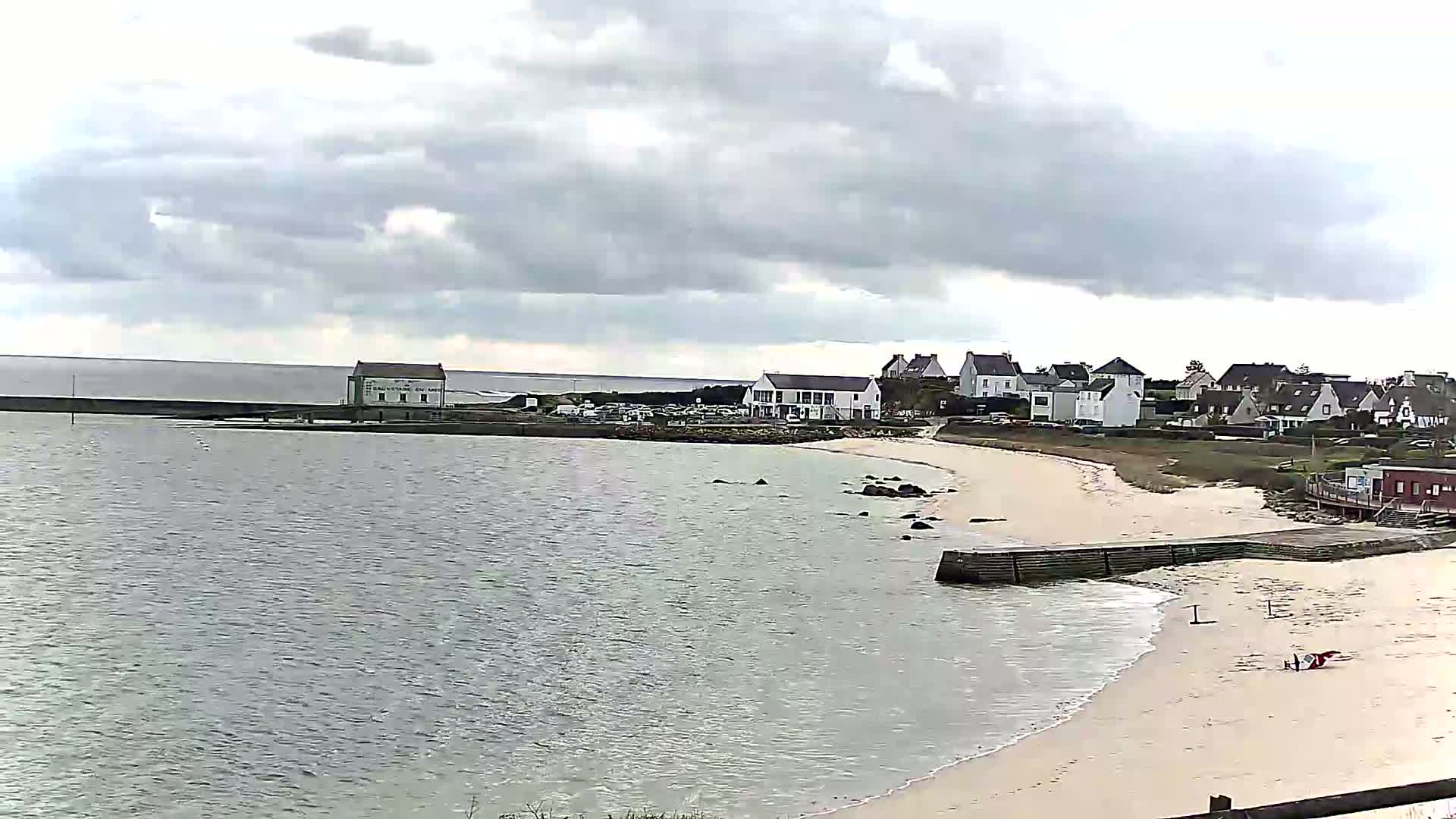 A calm body of water laps a sandy beach near a small coastal village under a cloudy sky.