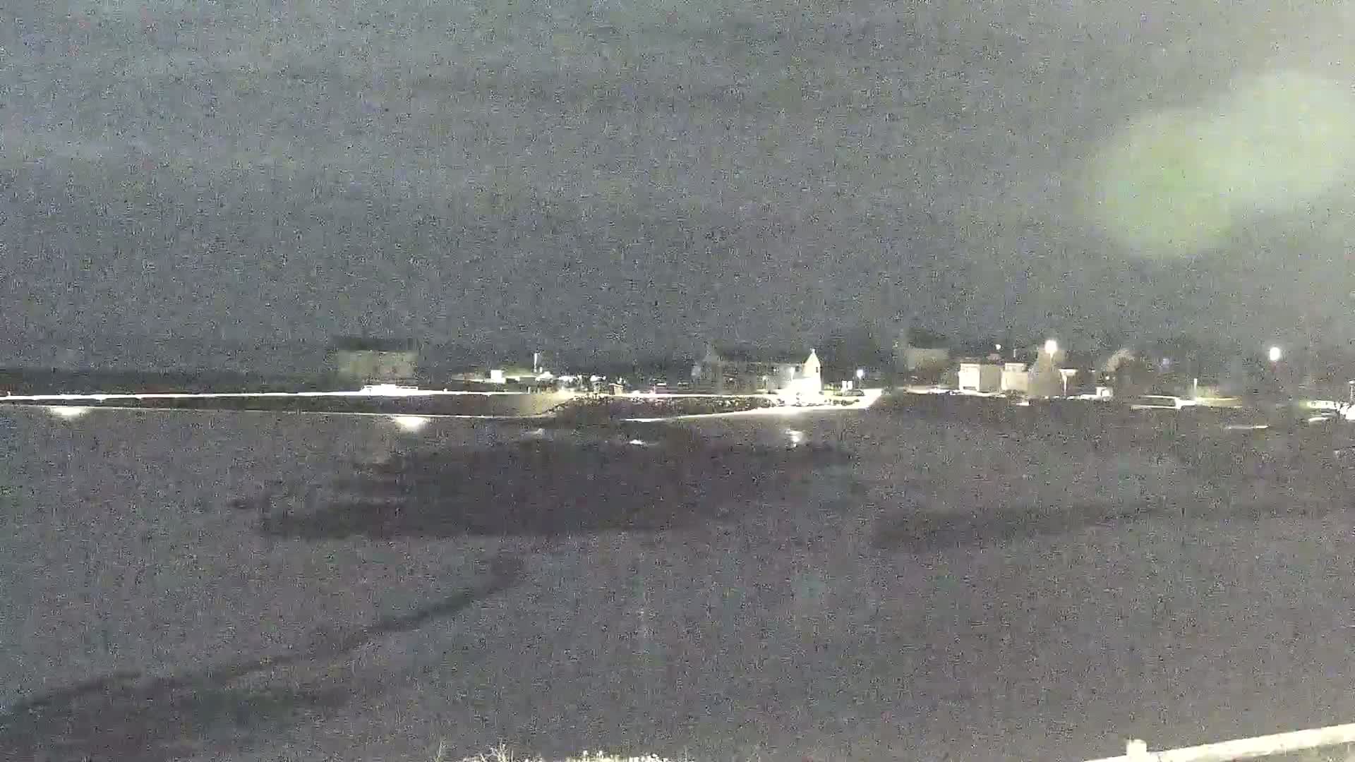 A nighttime, long shot of a waterfront town with faintly lit buildings under a dark, overcast sky.