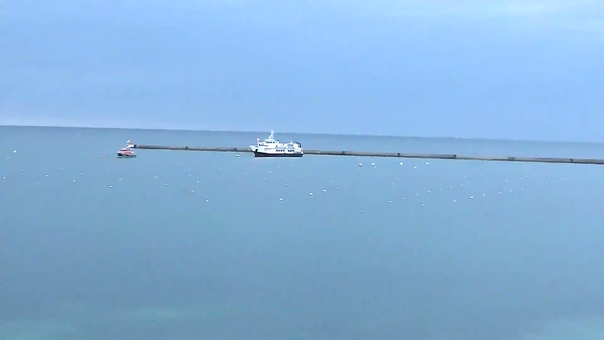 A ferry and a smaller boat are visible on a calm, light-blue sea, near a long, low breakwater.