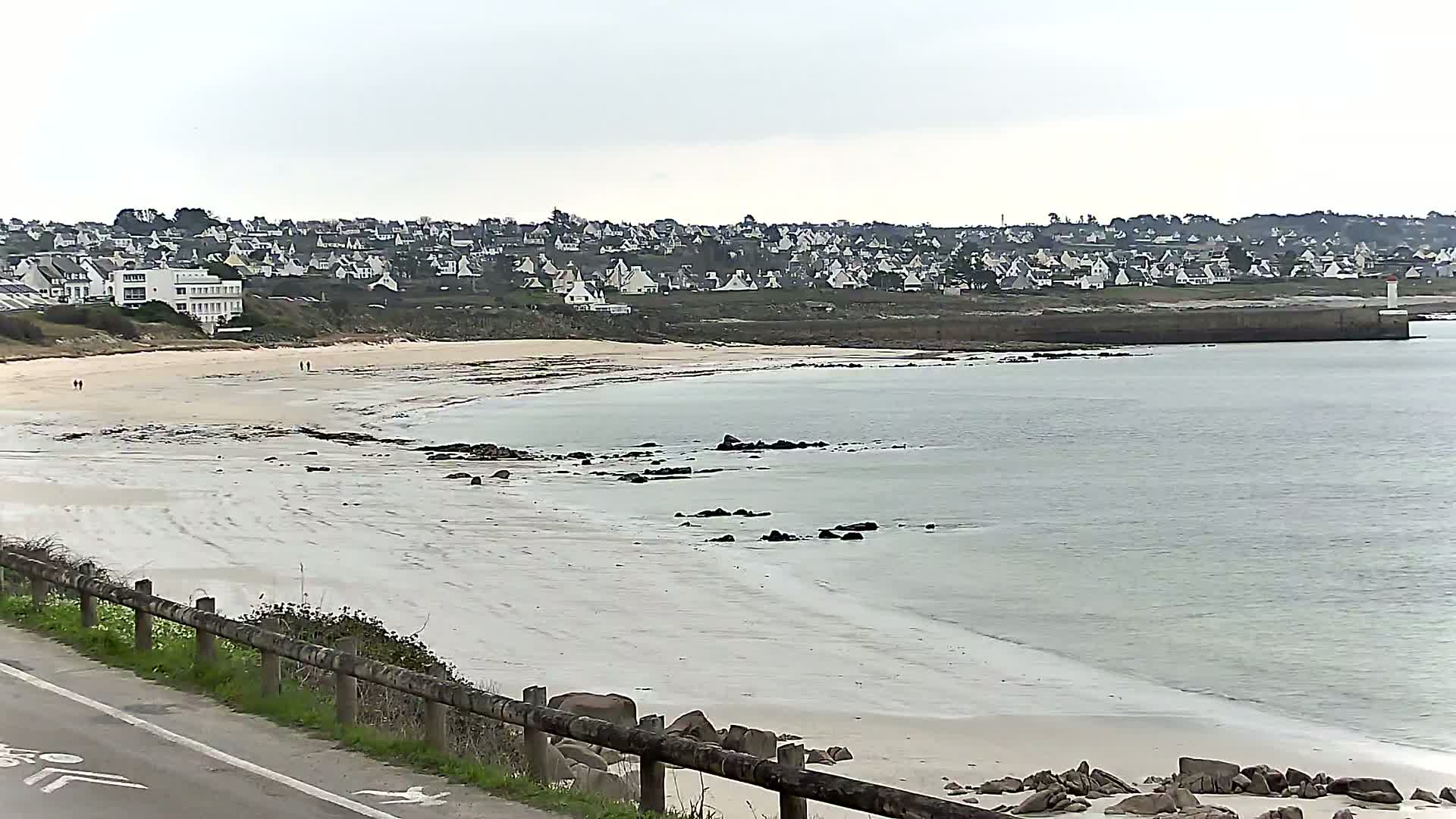A sandy beach with calm, light-grey water stretches below a hill dotted with white houses under an overcast sky.